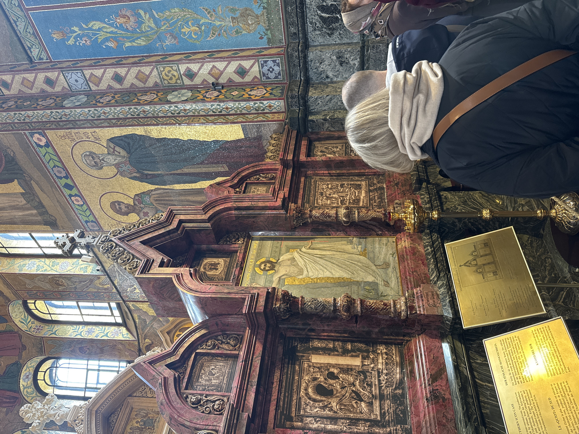 The image depicts the interior of the Church of the Savior on Spilled Blood in Saint Petersburg, Russia. The view is angled upwards, focusing on a portion of the elaborate marble and mosaic decorations adorning the walls and ceiling. A woman with blonde hair and wearing a dark jacket is looking at the displays. Various plaques in multiple languages including Spanish and English are visible. The plaques describes the icons and construction. A stained glass window provides natural light.