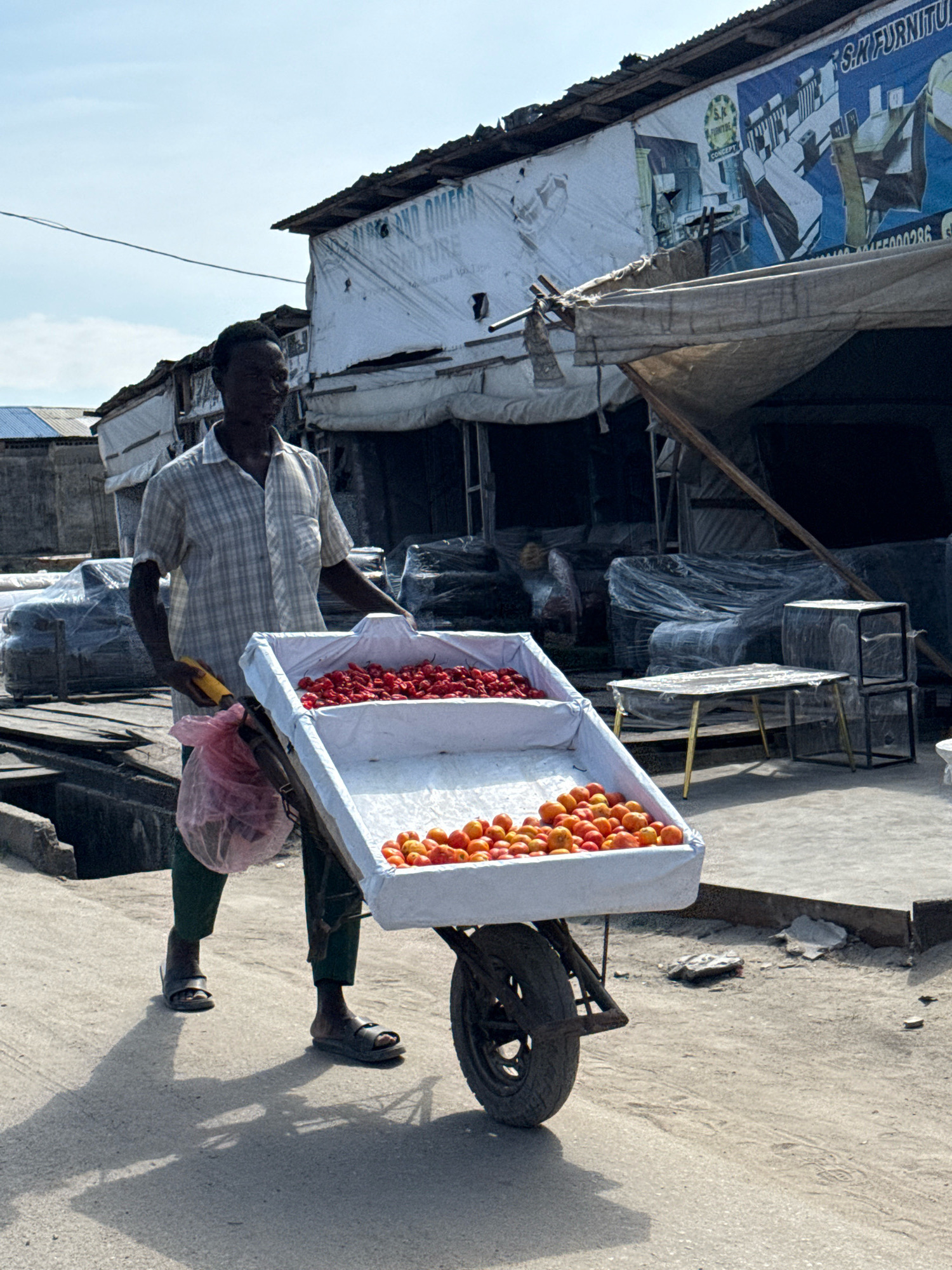 A full-shot, eye-level, outdoor photograph shows a Black man, wearing a checkered short-sleeved shirt and green pants, pushing a wheelbarrow filled with produce through the city of Ado, Nigeria. The man is walking on a paved ground, and his shadow is cast on the ground. The wheelbarrow, made of a white material, has two compartments; the top compartment is filled with small, red chili peppers, and the bottom compartment is filled with small, round, orange tomatoes. A pink plastic bag is hanging from the left handle of the wheelbarrow. In the background, a building with a corrugated iron roof is visible, with signs that read "S.M FURNITURE" and "ALPHA AND OMEGA FURNITURE". Furniture wrapped in plastic sheeting is stacked outside the building. The sky is clear and blue.