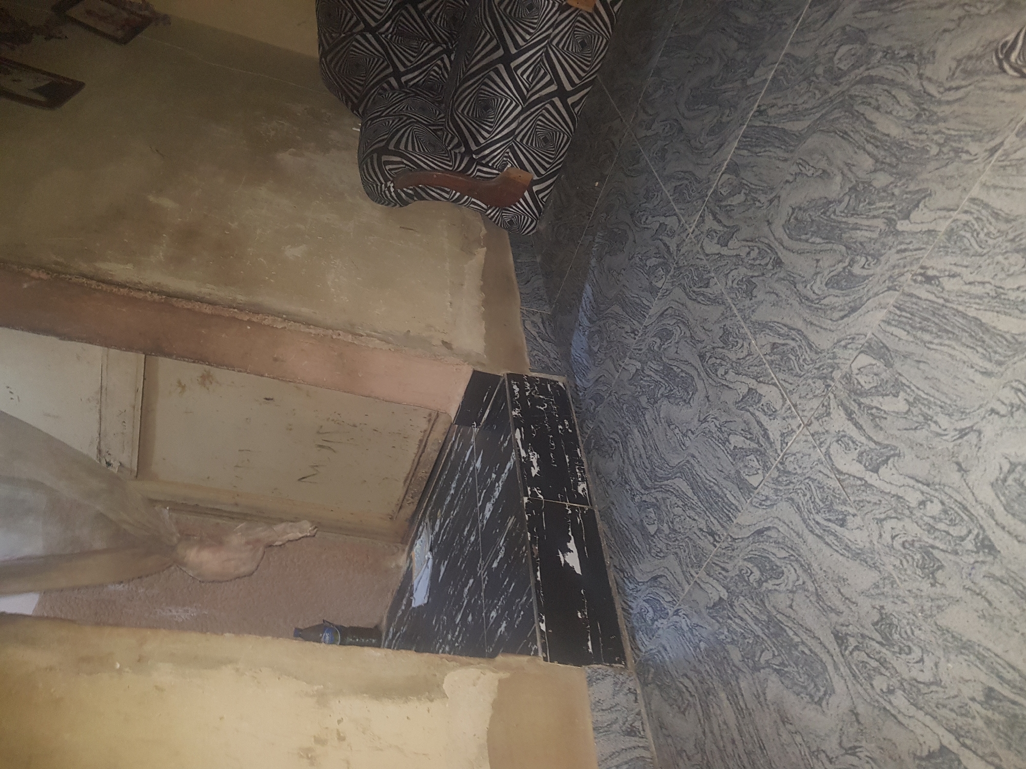 The image shows a view of an interior space in Hanwa, Nigeria. A person wearing patterned black and white pants is partially visible. The floor is tiled with a grey, swirled pattern. Adjacent to the tiled floor is a section of flooring covered in black and white tiles. A wall with tan paint is visible. There appears to be a metal pipe in the bottom left corner. The overall lighting is dim.