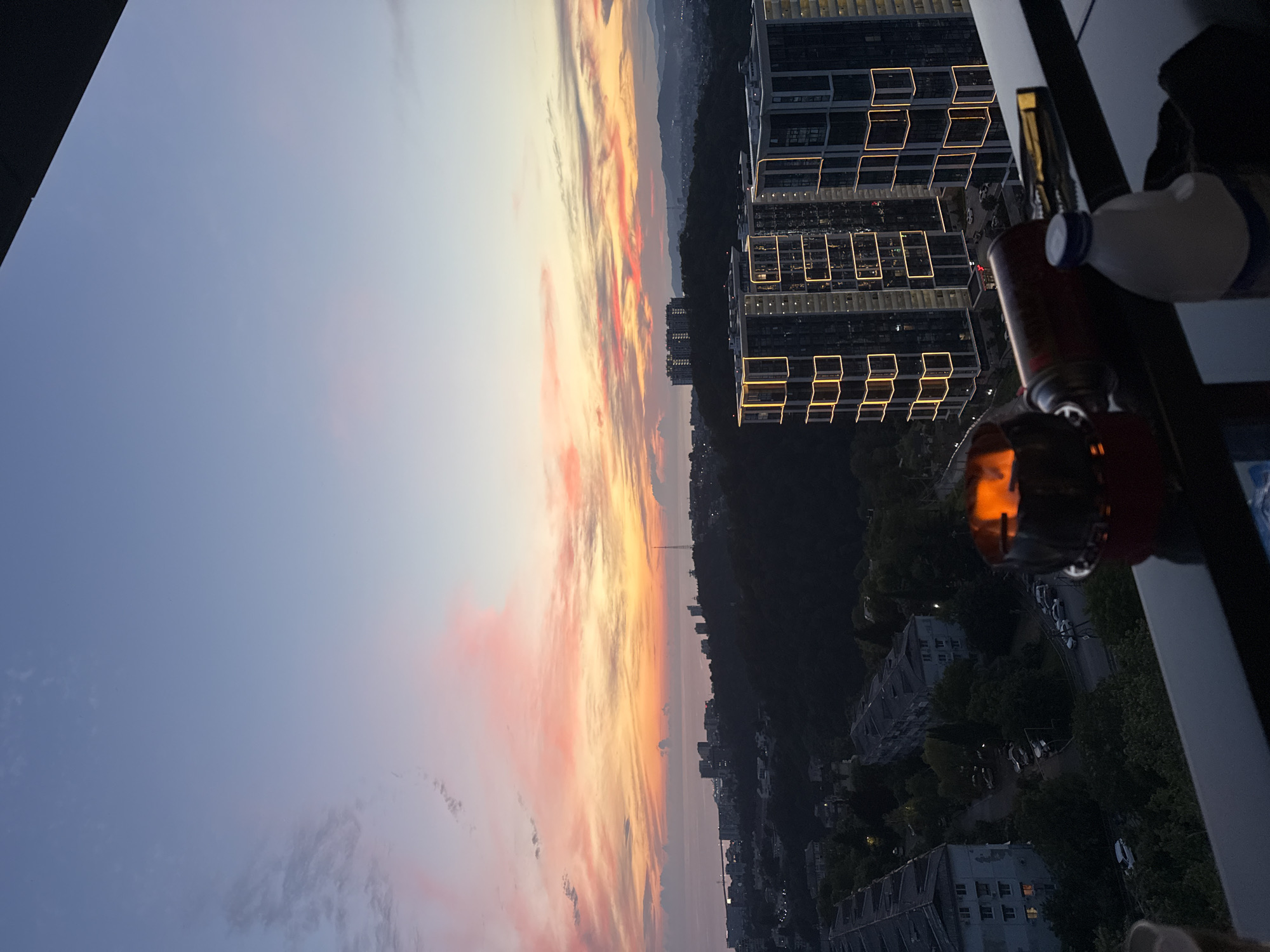 This image depicts a sunset over the city of Sochi, Russia, viewed from a balcony or elevated position. The sky is filled with dramatic clouds painted in hues of pink, orange, and yellow, transitioning to a pale blue at the top. In the foreground, on a dark railing, are a few objects, including a small portable grill with flames visible inside, a red gas canister, and a white plastic bottle with a blue cap.
The city of Sochi is visible in the mid-ground, with a dense collection of buildings, some appearing to be apartment complexes, nestled amongst dark green hills. Two modern high-rise buildings with illuminated yellow window frames stand out prominently. Further in the distance, the Black Sea can be seen, with a faint coastline and hills forming the horizon. The overall atmosphere is one of a peaceful evening winding down, with urban life set against a beautiful natural backdrop.