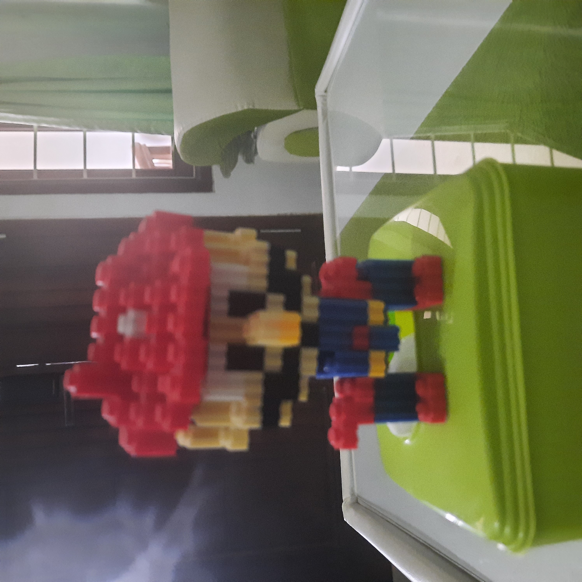 A pixelated, low-resolution image shows a toy figurine, resembling the character Mario, constructed from small, interlocking plastic bricks. The figurine is red, yellow, blue, and white, with a red hat. It stands on a bright green box, which is placed on a reflective surface that appears to be a table. In the background, there is a window with light green curtains and a glimpse of furniture, possibly a sofa, in a room. The overall impression is that of a domestic setting.
There is no indication that this image is located in the city of Indramayu, Indonesia.