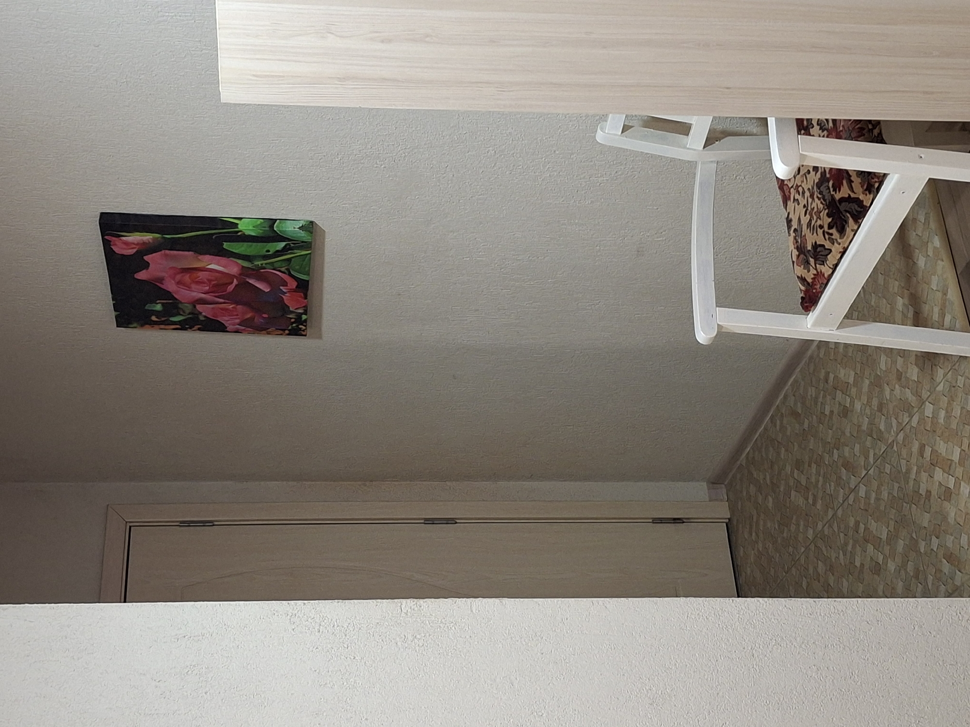 A tilted, eye-level shot captures an interior scene. A painting with pink roses is affixed to the white wall. A light-colored wooden table is partially visible at the top, with a white chair angled toward it. Part of a door with a light wood grain and a white wall below it are visible at the bottom. The background includes a tiled wall in shades of beige and brown. The location is an indoor setting in Krumovgrad, Bulgaria.