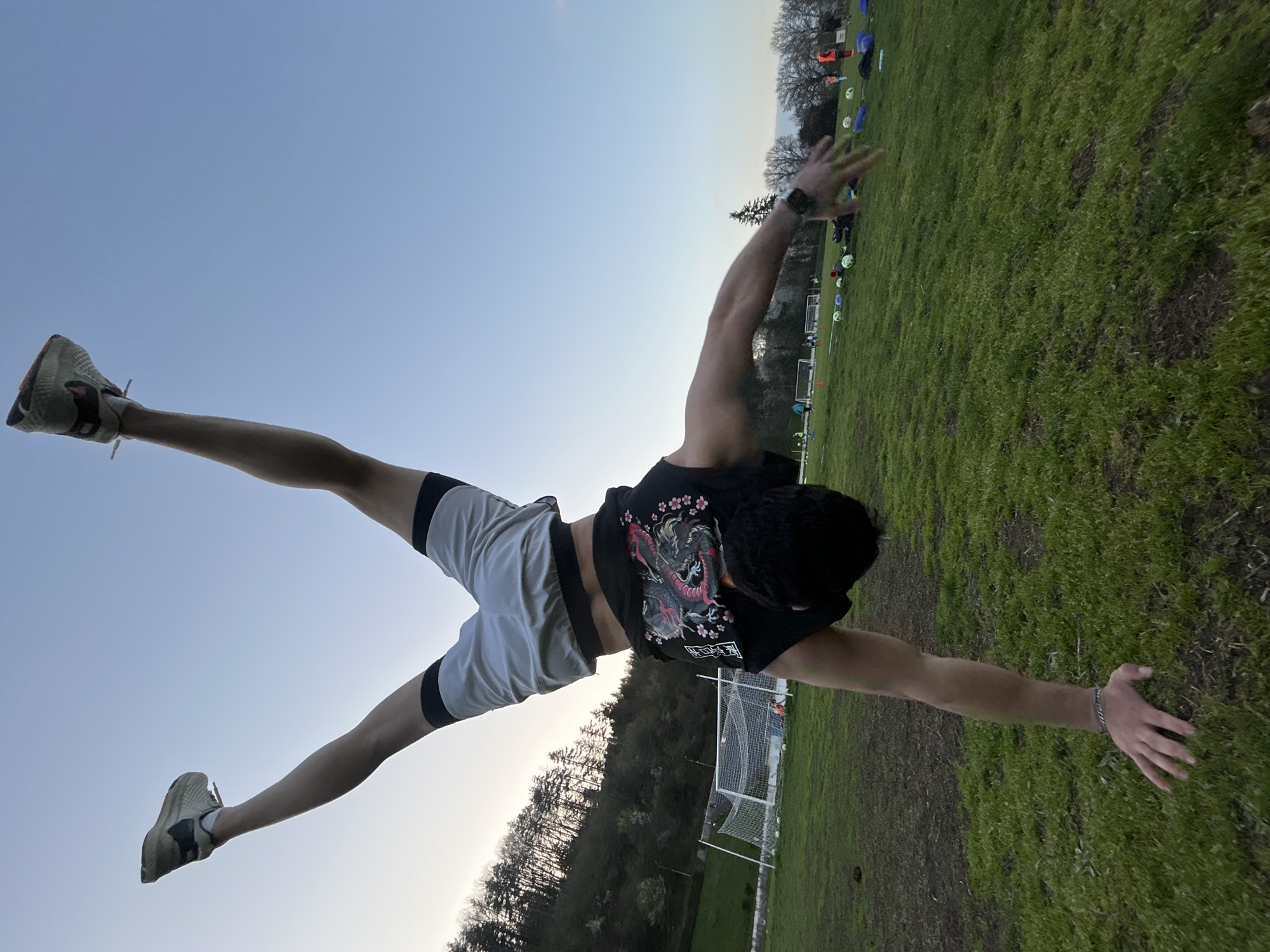A male is captured in a dynamic handstand pose with legs extended wide apart in what appears to be an outdoor field possibly in Troyan, Bulgaria. The man wears a black t-shirt featuring an image of a dragon and cherry blossoms on the back, paired with gray athletic shorts with black trim and tan sneakers. The man's wrists are adorned with a watch on his right and a bracelet on his left, and the surrounding environment suggests the man could be on a soccer field. A soccer net and training markers are in the background.