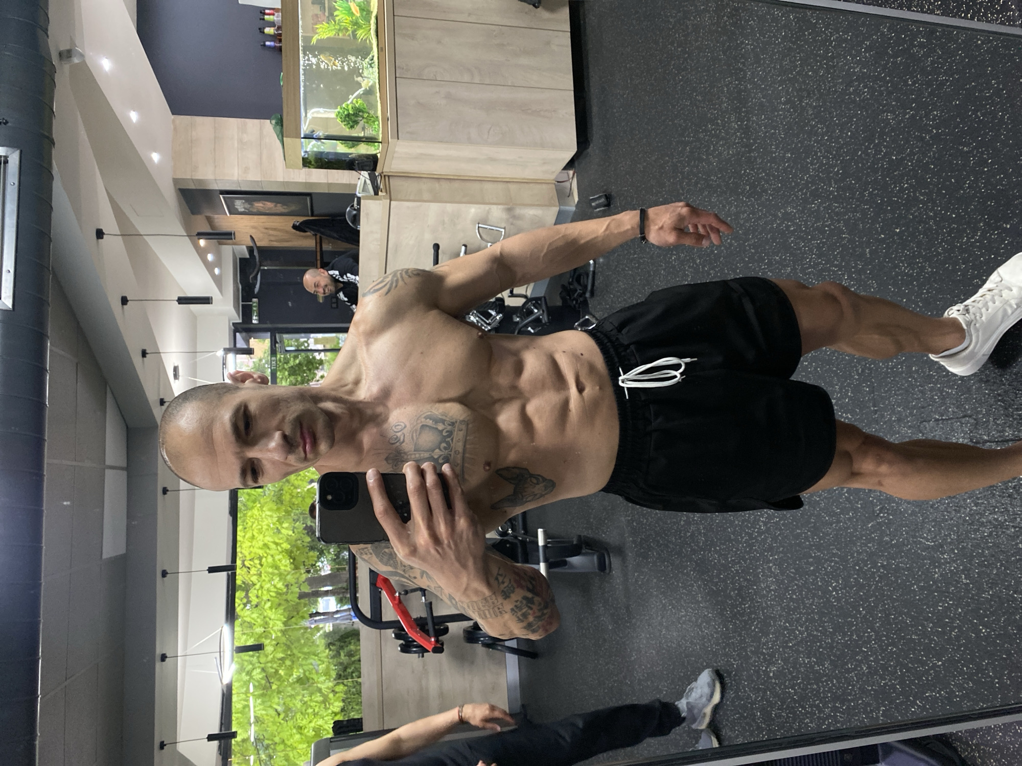 A man with tattoos on his upper body is taking a selfie in a gym. He is shirtless, wearing black shorts, and white sneakers. He is holding a smartphone in his right hand. The gym has black flooring, and there's weightlifting equipment visible in the background. A fish tank is located on the wall. A man in a suit sits on a chair. Reflection shows another individual partially. The selfie is taken from a mirror. The location may be Pleven, Bulgaria, based on the available information.