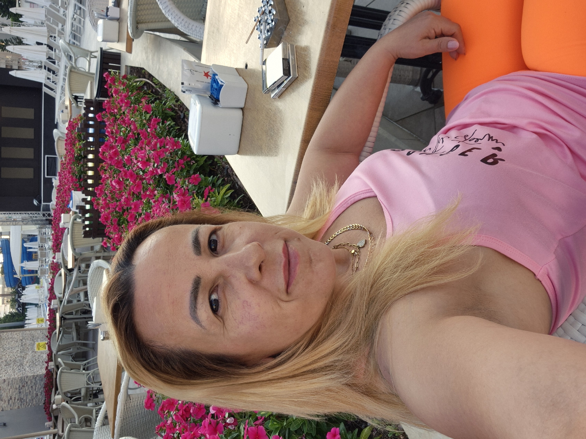 A woman with blonde hair and a pink tank top takes a selfie. She is sitting at a table outdoors, with pink flowers behind her and several white lounge chairs and umbrellas in the background. On the table, there is a pack of cigarettes, an ashtray with a lit cigarette, and a small white container.