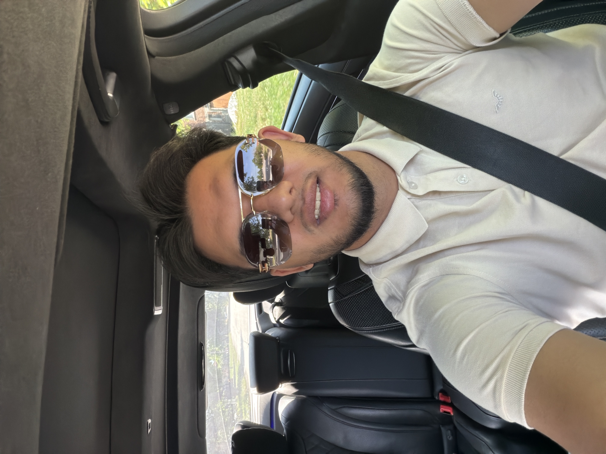 This image, set in Sinitovo, Bulgaria, captures a young man taking a selfie inside a car during what appears to be a bright, sunny day. The image is oriented with the man's head on the left and his body extending to the right, suggesting it was taken with the phone held vertically and then rotated.
The subject is a man in his 20s or early 30s with dark hair and a neatly trimmed beard or stubble. He wears stylish, aviator-style sunglasses with reflective lenses and gold-toned accents. His expression is neutral to slightly smiling. He is dressed in a light-colored, possibly beige or off-white, short-sleeved polo shirt, which features a small, embroidered emblem resembling a stylized sun or crown on the left chest. A black seatbelt is visible diagonally across his chest.
The scene is set within the interior of a car, which appears modern and well-maintained. The interior features dark-toned upholstery, likely black leather, with a visible quilted pattern on the rear seats, suggesting a premium vehicle. The car's headliner, A-pillar, and door panels are also dark-colored. Through the car windows, a clear, bright outdoor environment is visible, dominated by lush green foliage and hints of a building or structure in the background, indicative of a suburban or rural setting.
Notable details within the car interior include a white illuminated button or light on the roof console and a clearly visible "AIRBAG" label on the passenger side A-pillar. The overall impression is one of leisure, with the bright lighting suggesting mid-day.