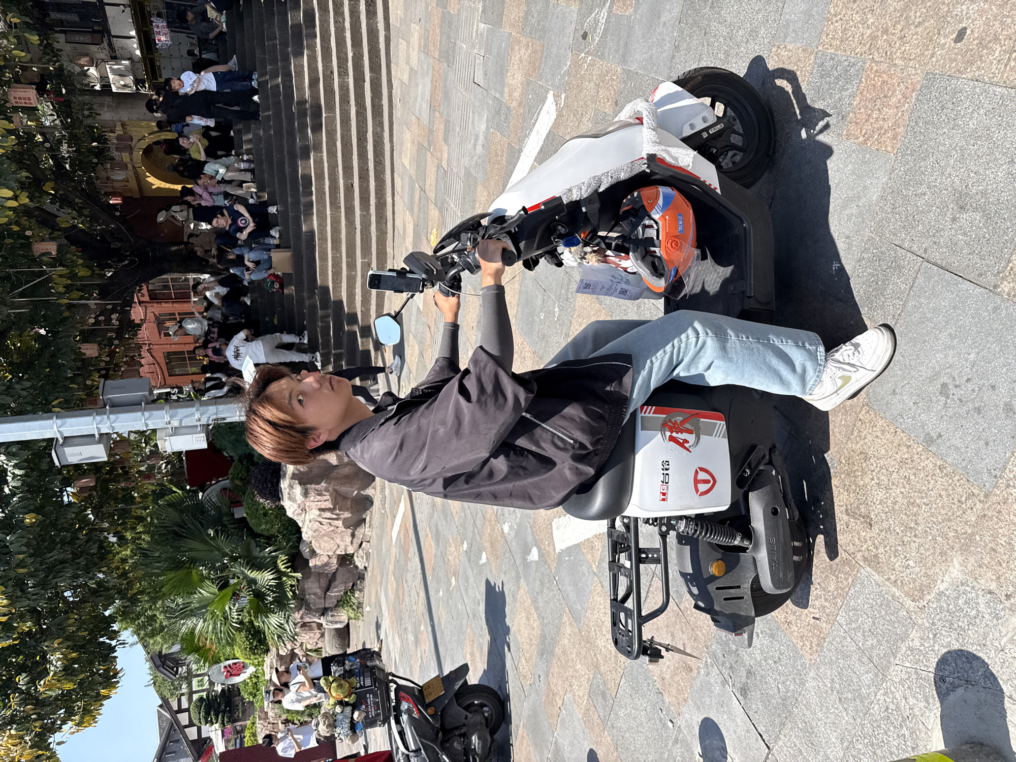 A medium, eye-level shot shows a person seated on a white, three-wheeled electric scooter, facing forward. The individual is clad in a black jacket, light blue jeans, and white sneakers, their hands on the scooter's handlebars. An orange and blue helmet rests on the scooter's front. The scooter has a black luggage rack.
The environment includes a patterned stone surface with an indistinct building and vegetation in the background, visible on the left. To the right, a set of stairs is partially visible with people sitting on them. Another parked scooter is visible in the lower left corner.
Based on landmarks and architectural style, the location is possibly in or near Ciqikou Ancient Town in Shapingba District, Chongqing, China.
