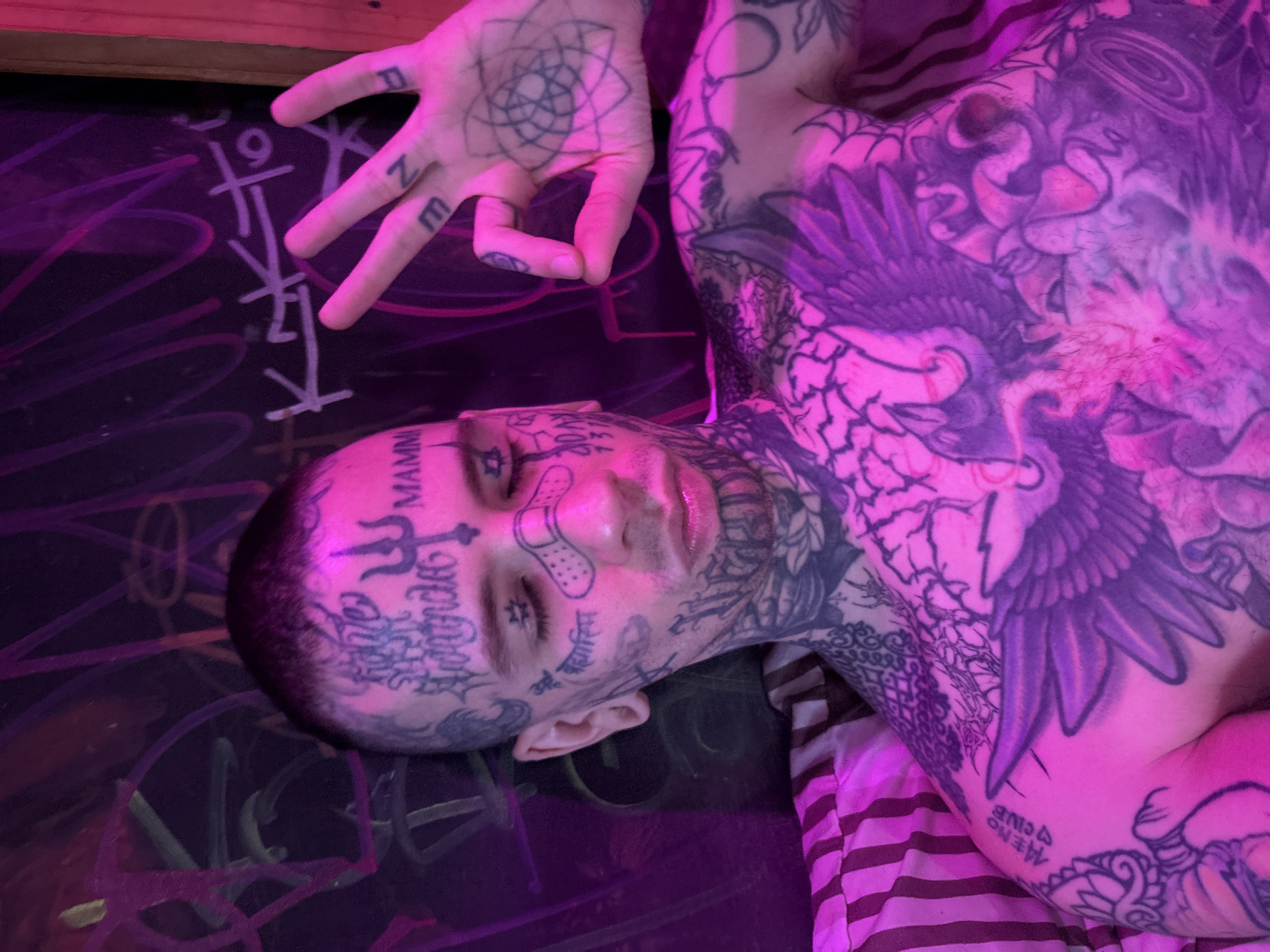 A man with extensive tattoos on his face, neck, chest, and arms is lying down on what appears to be a surface covered in graffiti, possibly a table or the ground. The man is fair-skinned and the tattoos are predominantly black ink. On his face, notable tattoos include symbols above and around his eyebrows, script across his forehead, and designs near his eyes and temples. He also has a bandage on his nose. His chest features a large tattoo, possibly of a bird of prey with outstretched wings. He has tattoos on his fingers as well. The graffiti on the surface contains various symbols and what may be writing. The lighting is primarily pink and purple. There is a striped fabric under his torso, which may be clothing or a bedspread.
There is no information available to me to determine if this image was taken in Buenos Aires, Argentina.