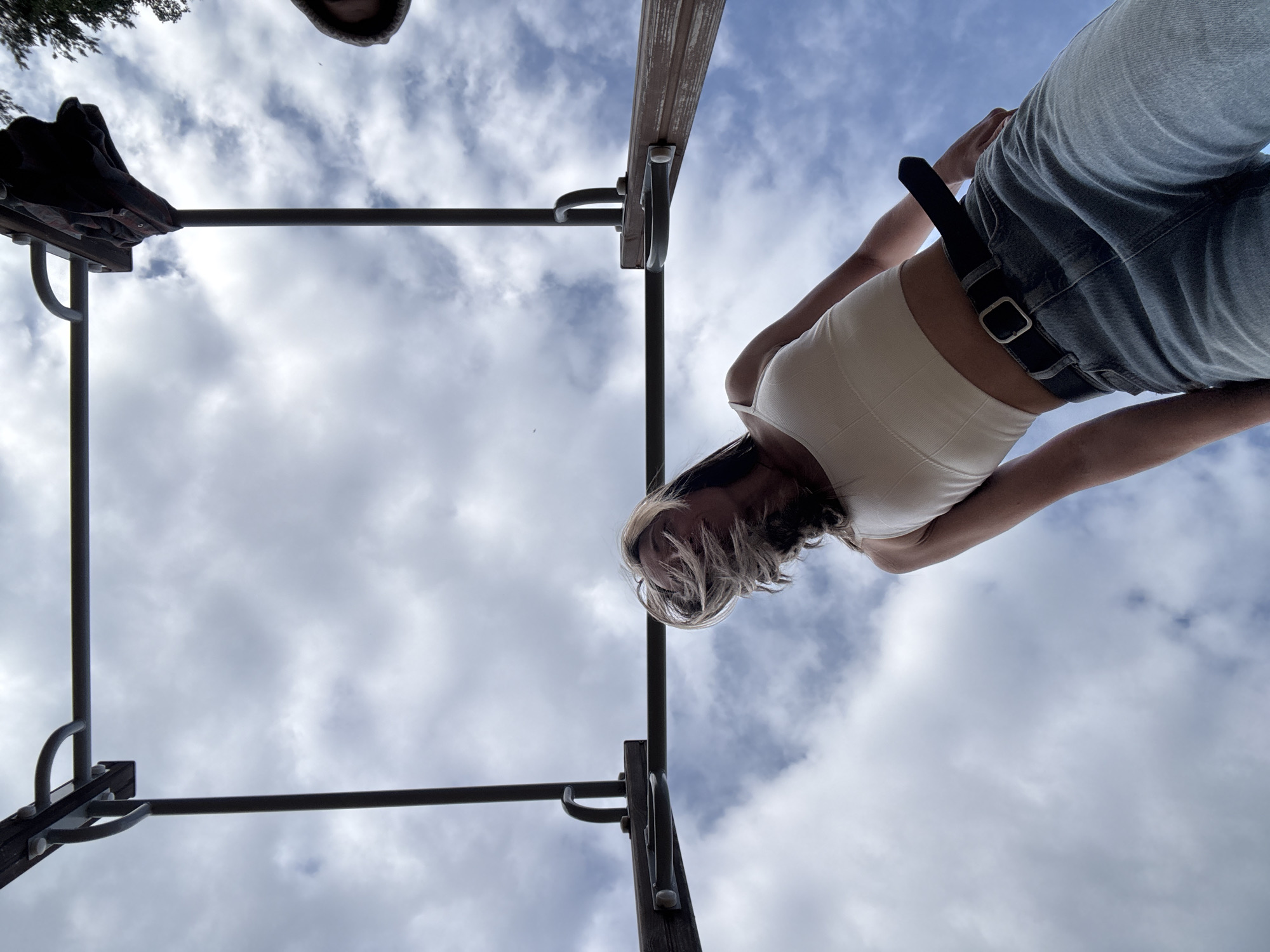 The image is a low-angle shot looking up at a woman standing on a metal climbing frame against a cloudy sky. The woman is wearing a white tank top and blue jeans with a black belt. Part of a dark-colored jacket and a hooded individual are visible at the top left corner of the frame. The location is unspecified.