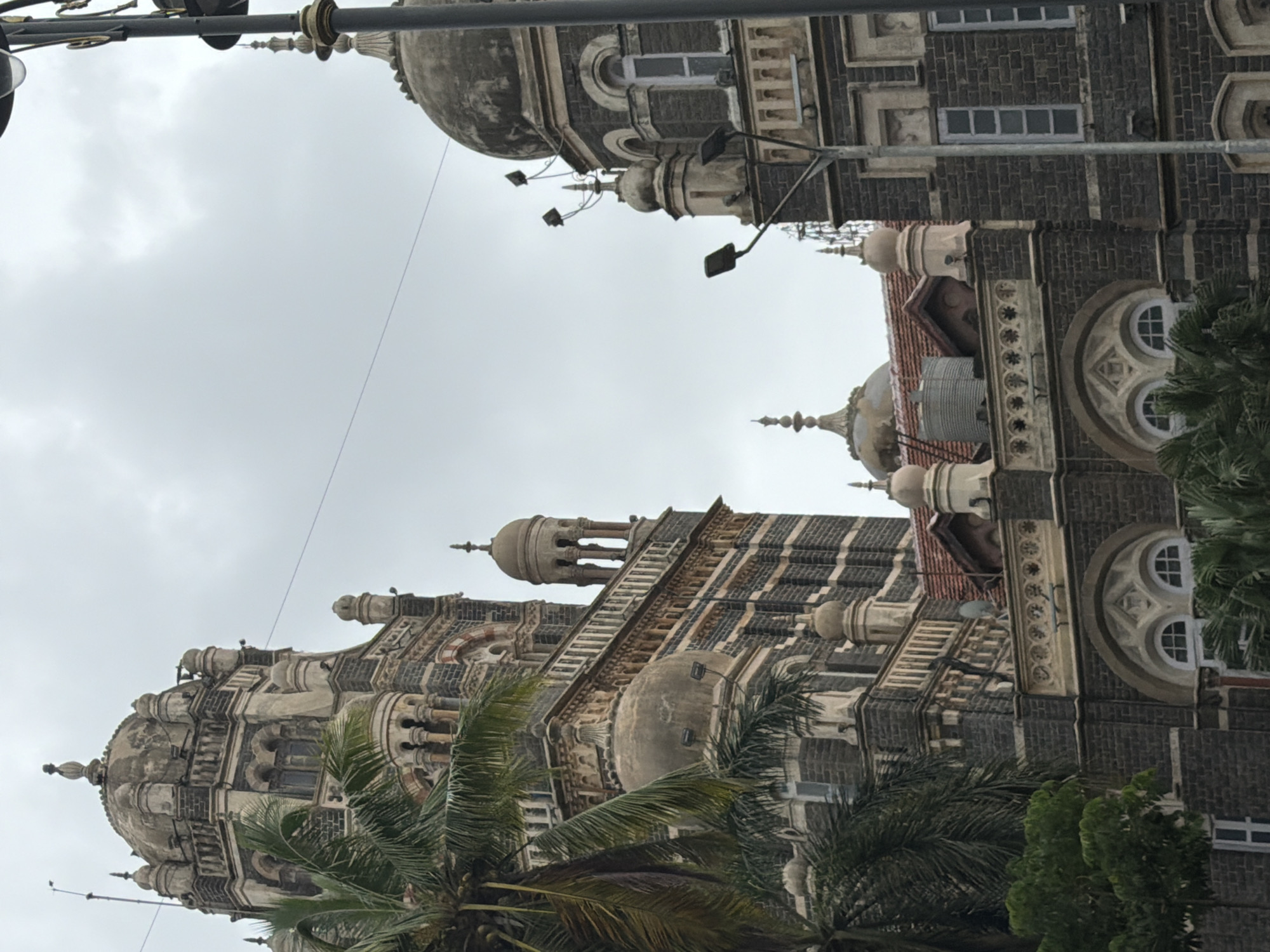 The image shows a close-up view of the Victoria Terminus station, now known as Chhatrapati Shivaji Terminus, in Mumbai, India. The architectural style is a blend of Victorian Gothic Revival, Indian, and Islamic influences, characterized by its ornate stonework, domes, spires, and arched windows. Two towers of the station are visible, one on the left and another to the right, partially obscured by palm trees and a lamppost. The sky is overcast and gray, suggesting a cloudy day. The building's facade is constructed from dark, textured stone, with lighter stone detailing around windows and along decorative bands. Several arched windows with white frames are visible, as well as smaller domed structures and pinnacles. A water tank is perched on one of the lower roofs on the right side of the image. The overall impression is of a grand and historic railway station.