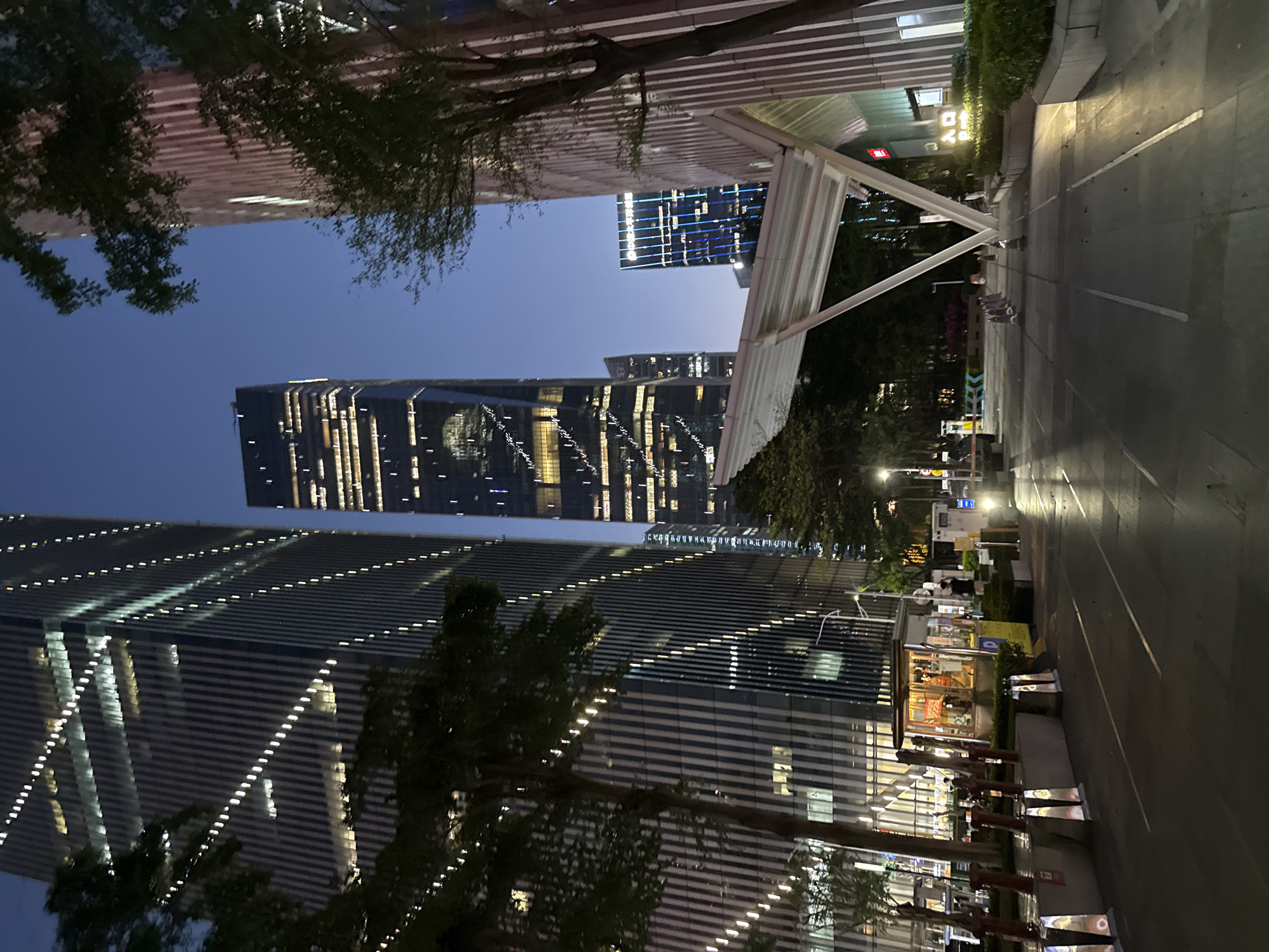 This is an evening scene in the Tianhe District of Guangzhou, China, featuring modern architecture and urban infrastructure. Two skyscrapers, illuminated with strings of lights and window patterns, dominate the view. A third, taller building with more dispersed lighting is visible in the distance. The foreground shows a well-lit pedestrian area with trees and outdoor seating or kiosk structures, and a covered walkway. No people are clearly visible. The environment is modern and urban, showcasing the contemporary architecture of the Tianhe District.