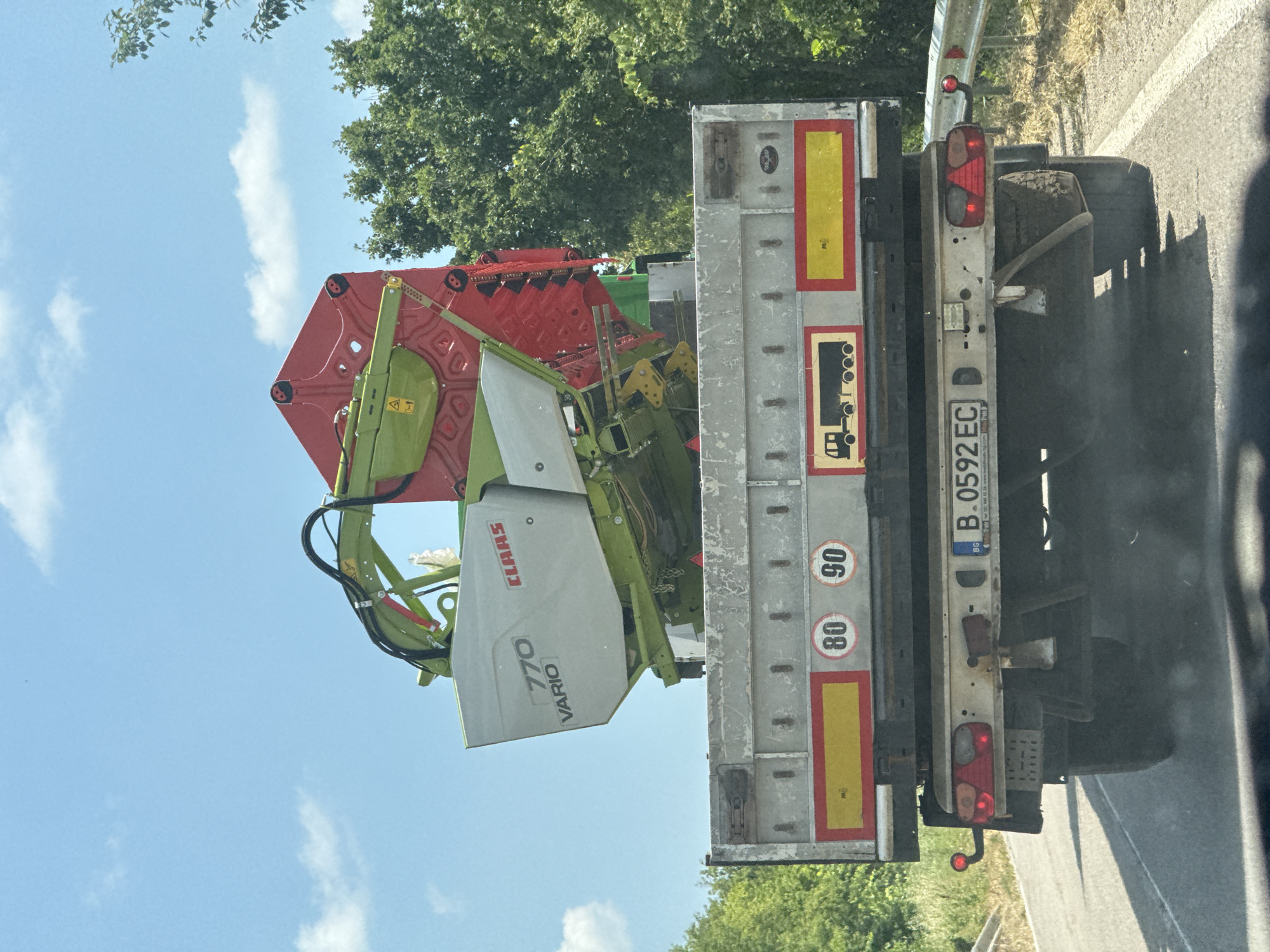 The image shows a close-up view of a trailer truck carrying agricultural machinery. The machinery, primarily green and red, appears to be a CLAAS 770 VARIO combine harvester. The truck is parked on the side of a road, with a highway barrier visible in the background, along with trees and a blue sky with some clouds. The time of day seems to be daytime, likely late morning or afternoon, given the bright sunlight casting shadows. The weather appears to be clear and sunny.
Visible text on the machinery includes "CLAAS" and "770 VARIO". On the back of the trailer, there are several safety markers, including speed limit signs (80 and 90) and reflective tape. A license plate is also visible, reading "B.0592 EC" with "BG" indicating Bulgaria. The truck itself is a flatbed trailer. The overall scene suggests the transport of agricultural equipment, possibly to or from a farm or for maintenance. The image is taken from a slightly angled perspective, with the truck positioned towards the right side of the frame. The condition of the machinery appears used, with some dirt and dust visible, which is common for agricultural equipment.