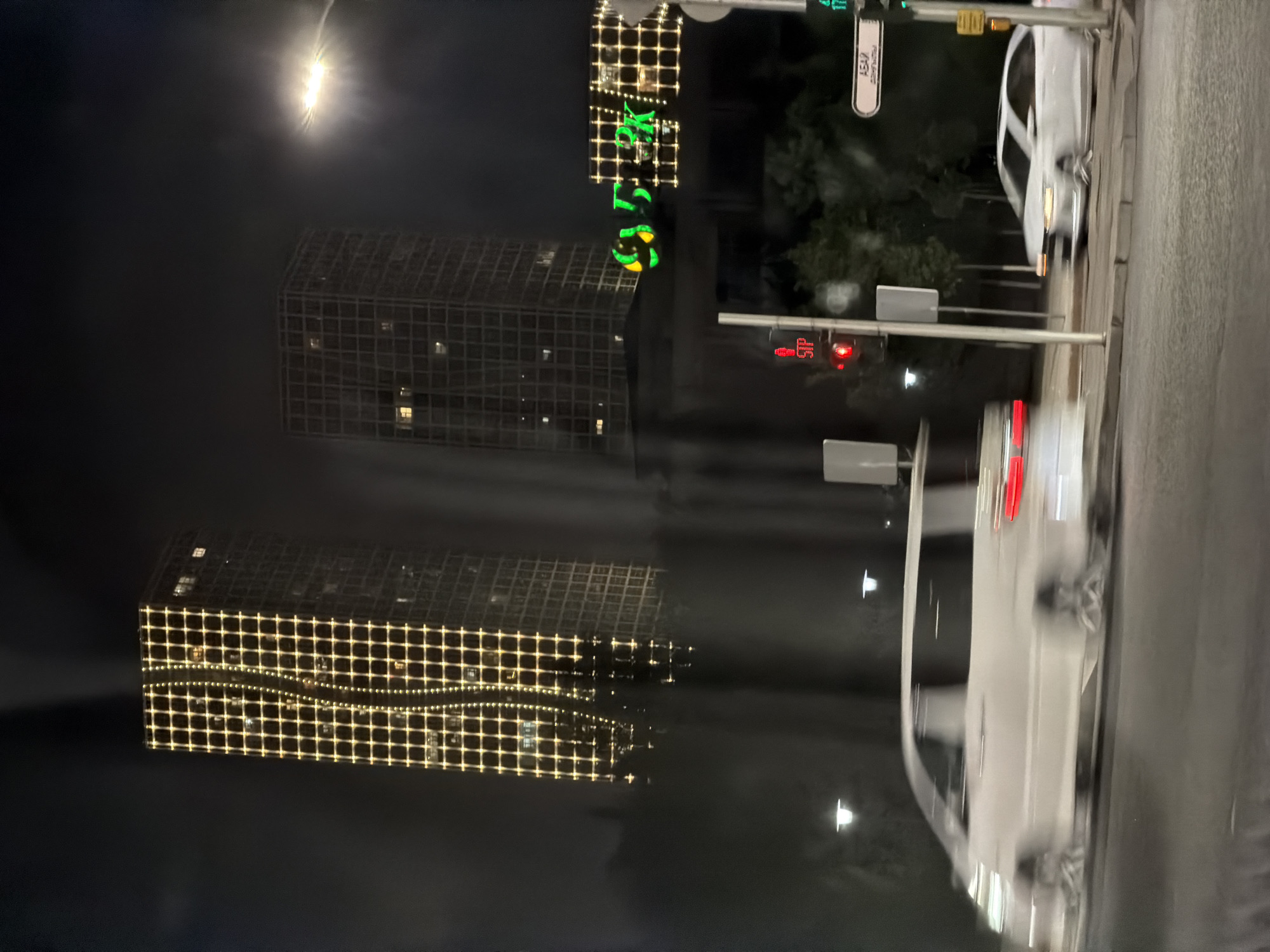 This image captures a night-time street scene in Astana, Kazakhstan, likely on a main thoroughfare. Two prominent high-rise buildings are visible in the background, their facades adorned with grid-like patterns of warm yellow lights. One building features a distinct vertical wavy line of smaller lights running down its center, creating a decorative effect. To the right of these buildings, a large, illuminated green sign with yellow accents displays text, though it is partially obscured and blurry.
In the foreground, the scene is dominated by a busy intersection. A white minivan is in motion, captured with motion blur, indicating it is either driving or turning. A white sedan is also present, moving on the right side of the road. Traffic lights are visible, with a red light illuminated on one pole and a green light on another. Street signs are also present, one with text that appears to be in a Cyrillic script. The street is paved, and the overall environment suggests an urban setting with street lighting and surrounding greenery, though the darkness and motion blur obscure finer details. The image seems to be taken from a moving vehicle, contributing to the overall blurriness.