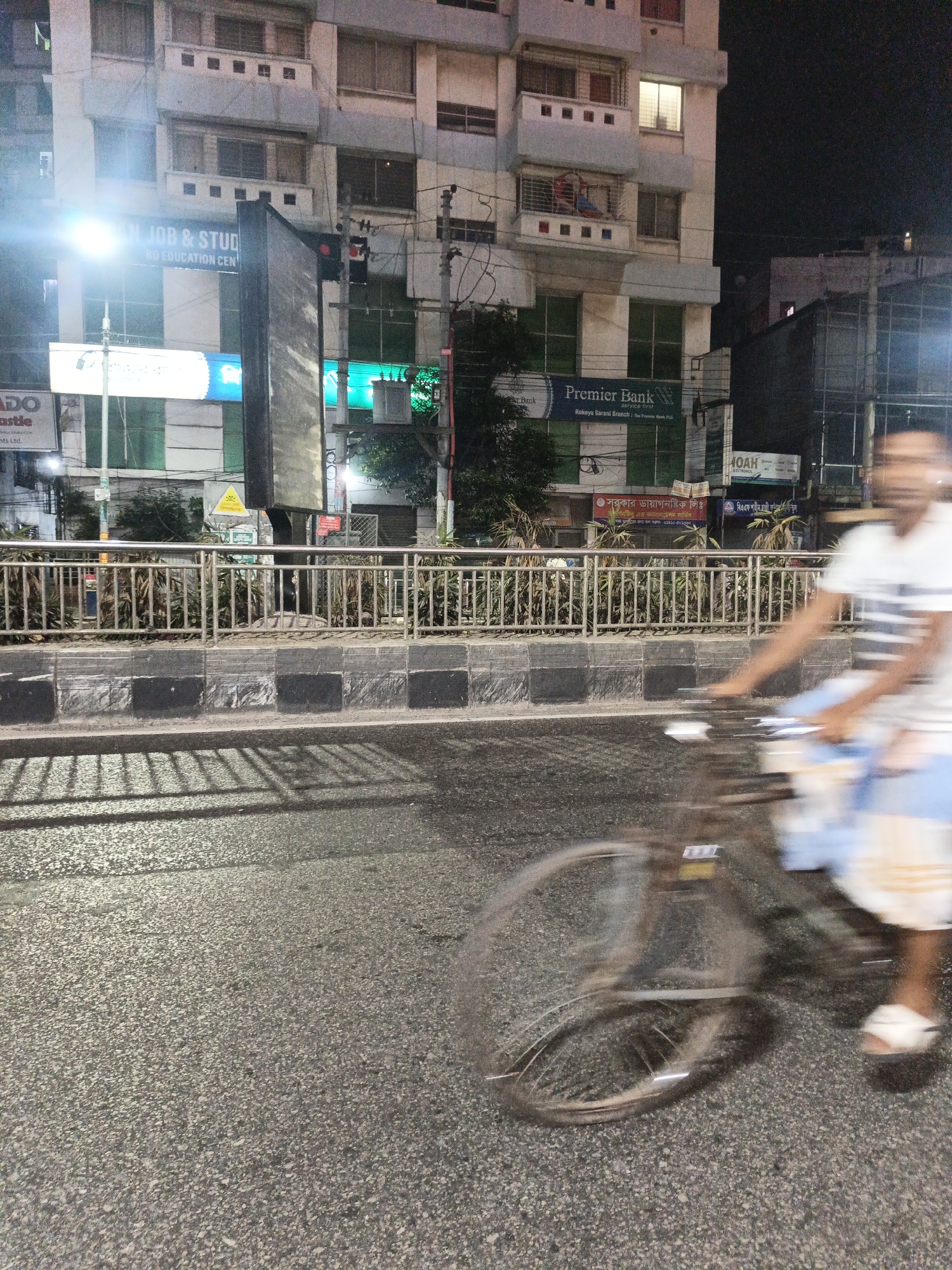 A nighttime street scene in Dhaka, Bangladesh, captures a person blurred by motion as they cycle past a building. The building's facade, illuminated by streetlights, displays various signs including "JOB & STUD BD EDUCATION CEN," "Premier Bank," and "NOAH ELECTRONICS." The foreground features a paved road and a metal railing separating the street from a sidewalk lined with greenery. The lighting suggests it is after dusk, with artificial light sources casting shadows.