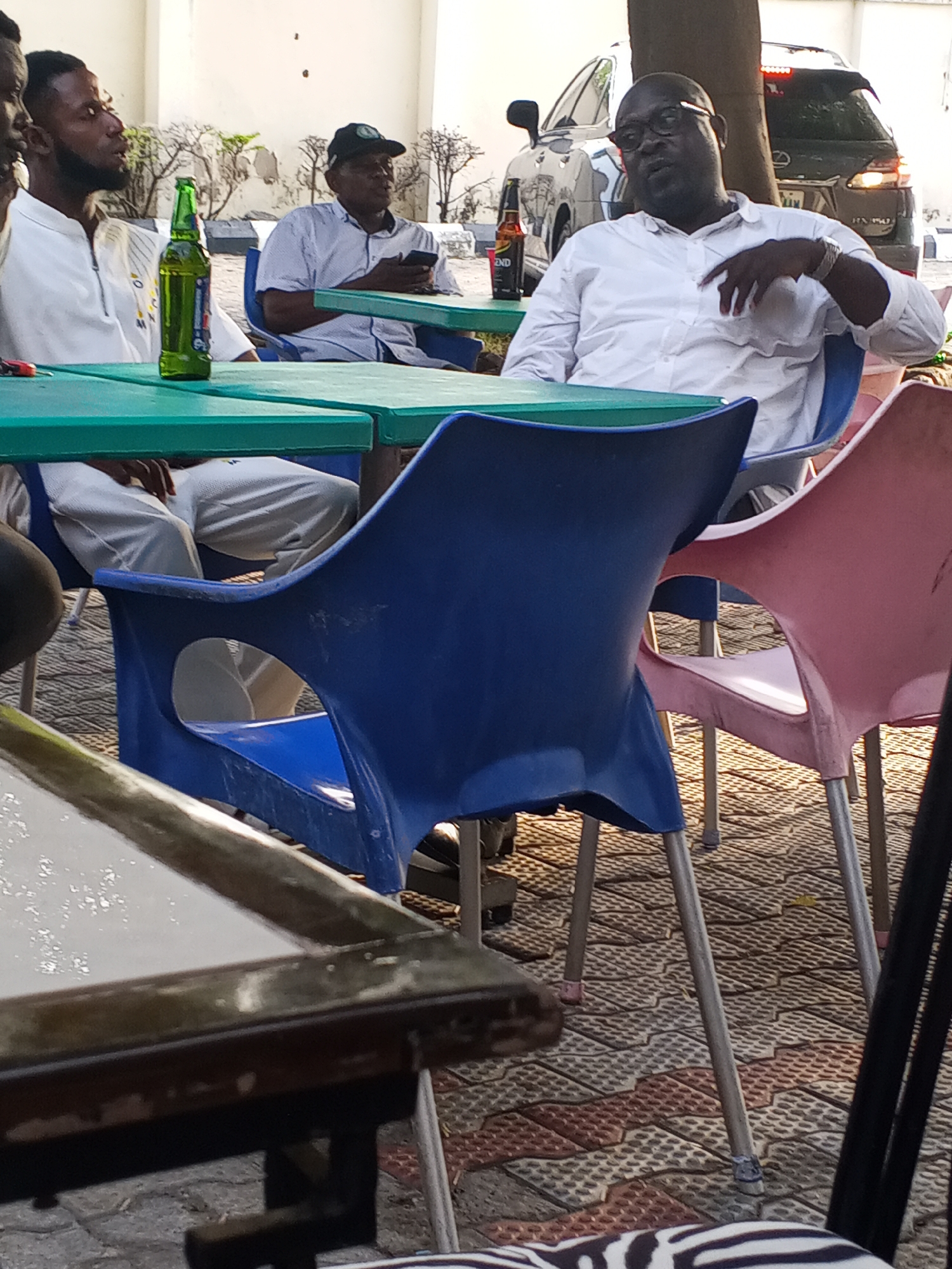 The image shows a group of four men sitting at plastic tables in what appears to be an outdoor setting. The location is most likely Lokoja, Nigeria.
One man, wearing a white shirt and glasses, is seated in a blue plastic chair at a table with a bottle of beer. He is gesturing with his right hand.
A second man in a light-colored shirt and a baseball cap is looking at his mobile phone and sitting at the same table. A third man, partially visible, is wearing a white top and sitting nearby.
In the foreground, there is a partially visible table with a dark frame, and a zebra-striped chair.
The scene suggests a casual social gathering in an outdoor seating area. A car, possibly a Lexus SUV, is parked in the background. The pavement is patterned with dark red and light brown bricks. A green bottle, assumed to be beer, is on the table. Another brown beer bottle is also present. The overall setting gives the impression of an outdoor establishment, possibly a bar or restaurant.