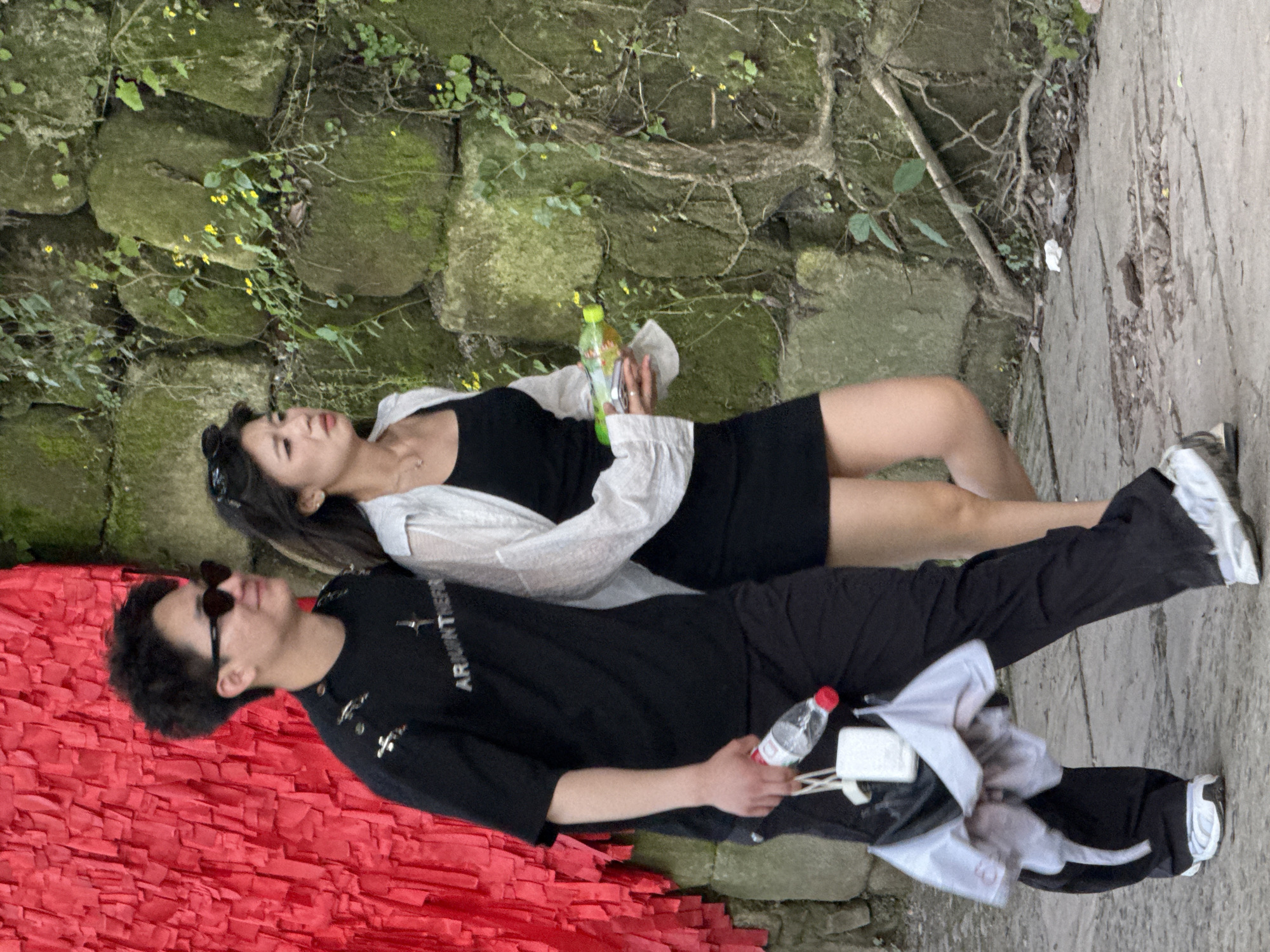Two individuals are lying down outside on a partially red surface, next to a stone wall covered with green vegetation and a concrete path. The woman is wearing a black dress and a white shirt, and has sunglasses on her head. She is holding a green and yellow drink. The man is wearing a black shirt, black pants, and sunglasses. He is holding a bottle with a red cap. The scene appears to be outdoors. The location is likely Beibei, China.