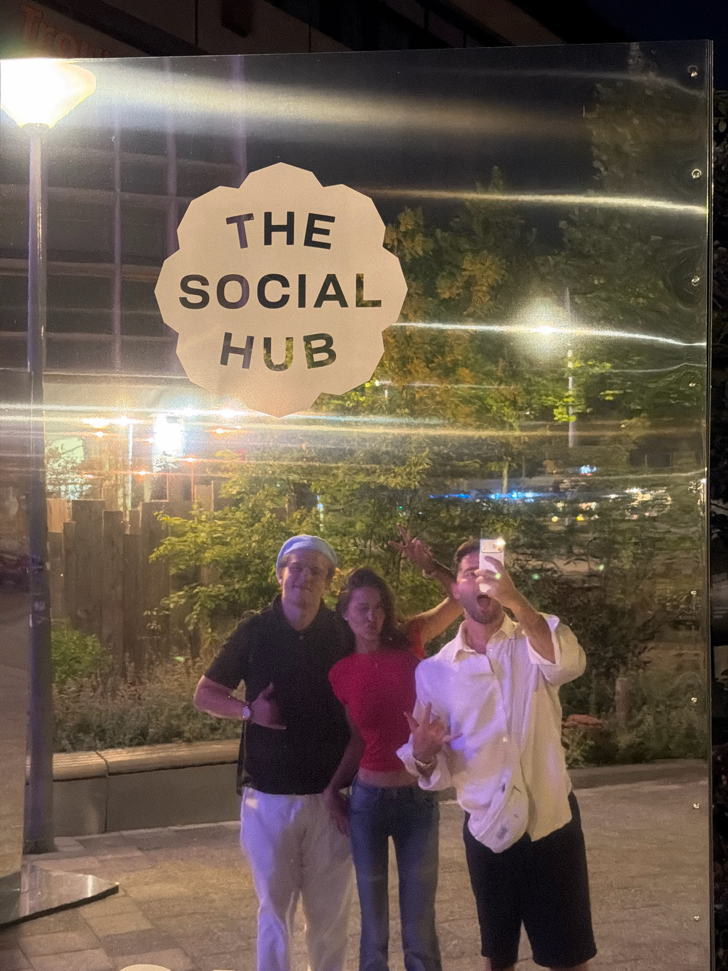 The image shows a reflective surface with text that reads "THE SOCIAL HUB" printed on a light-colored cloud-shaped decal. Three individuals are reflected in the surface, appearing to take a selfie. The person on the left is wearing a black polo shirt and white pants. The person in the middle is wearing a red top and blue jeans. The person on the right is wearing a white collared shirt and black shorts, holding up a smartphone to take the picture and making a peace sign. They are in an outdoor urban environment with trees and city lights visible in the background reflections. The overall lighting suggests it is nighttime or dusk.