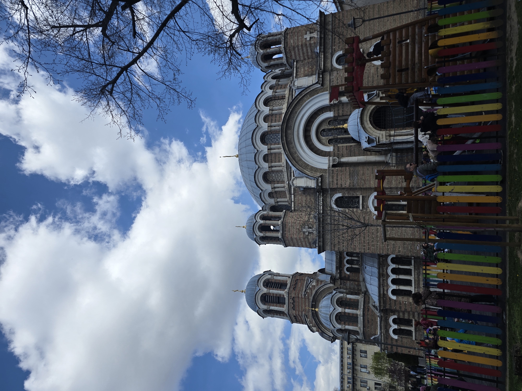The image shows the St. Sophia Church in Sofia, Bulgaria. The church is a Byzantine-style brick building with multiple domes and arched windows. Part of the church is obscured by a children's playground with brightly colored wooden slats. Bare tree branches are visible in the upper left corner, contrasting with the blue sky dotted with white clouds. Several people, including children, are interacting with the playground equipment. An apartment building is partially visible at the bottom.