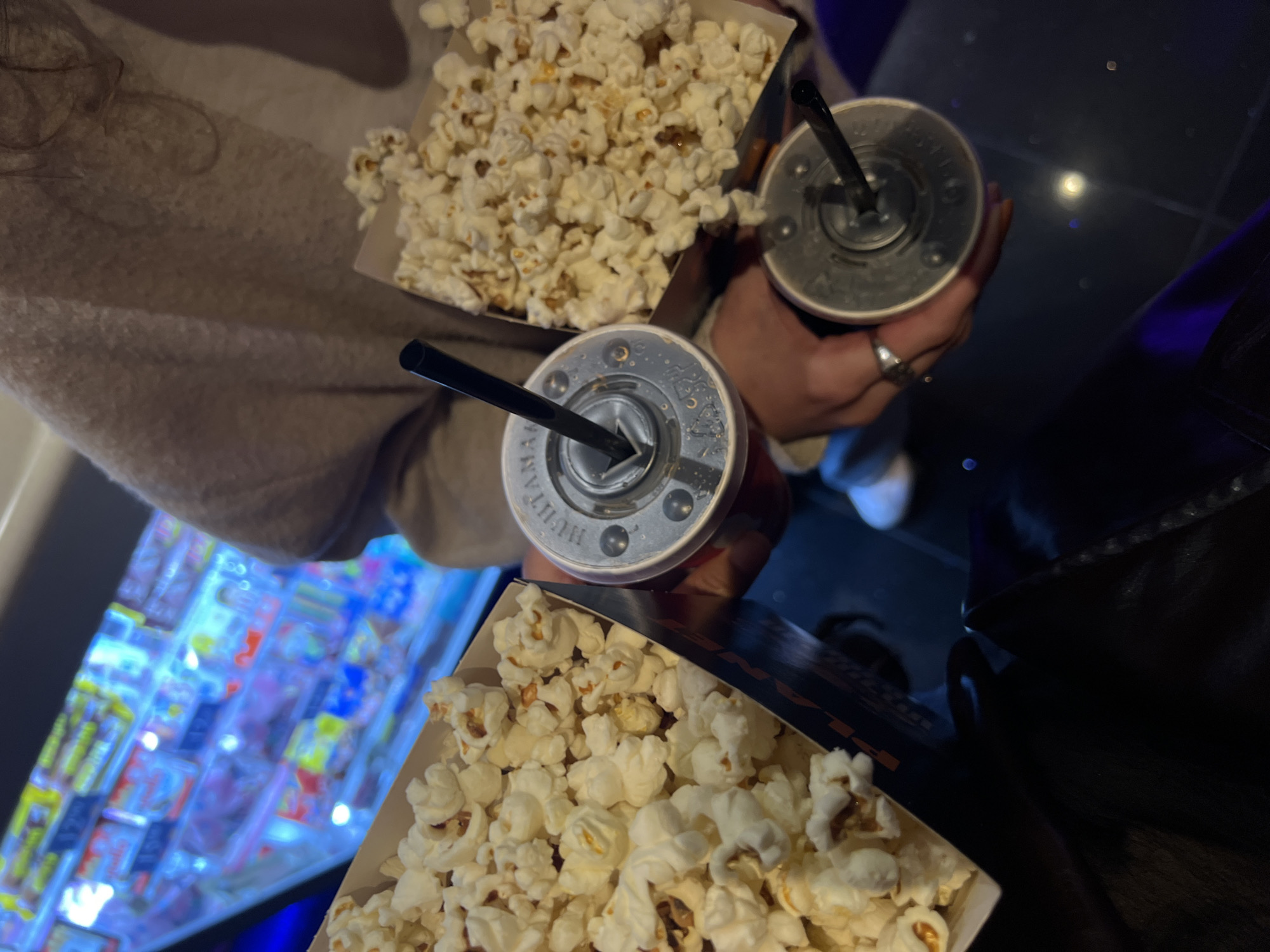 The image shows two individuals, seemingly at a movie theater in Rishon LeZion, Israel. Each person has a container of popcorn and a drink with a straw. The background includes what appears to be a vending machine with brightly colored items. The scene is dimly lit.