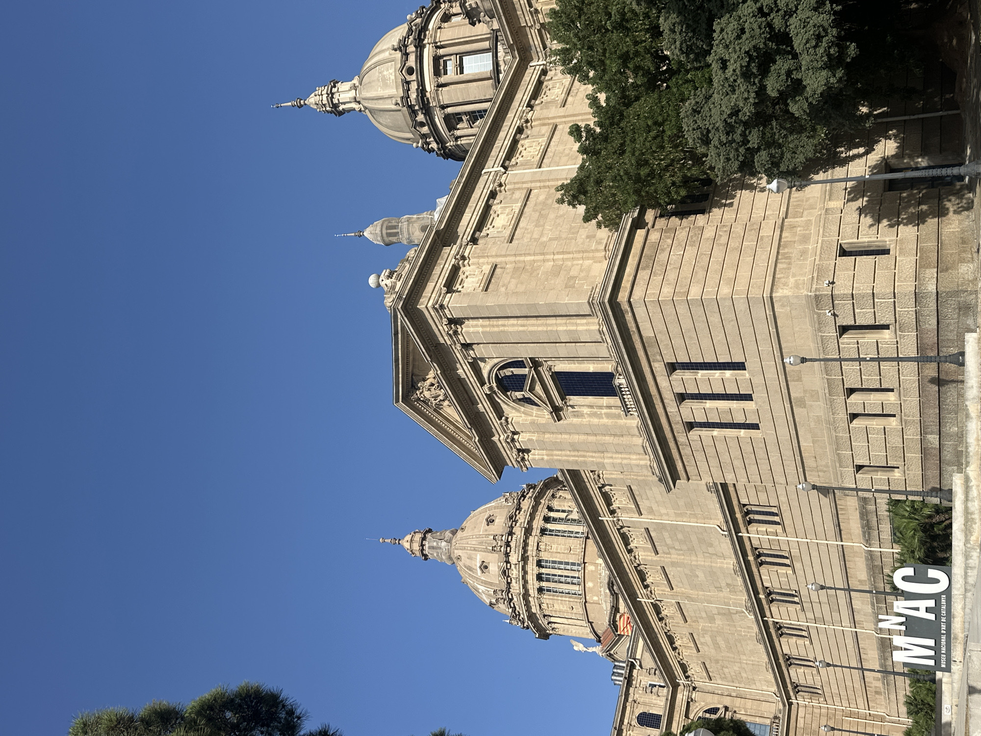 The image shows the Palau Nacional, the former Palace of National Art, now housing the Museu Nacional d'Art de Catalunya (MNAC) in Barcelona, Spain. The architecture is grand, with multiple domes and classical detailing, built with light-colored stone. The building is partially obscured by lush green trees. In the foreground, a large white sign with the letters "MNAC" is visible, indicating the museum's name. The sky above is a clear, deep blue. There are no people visible in the frame, and the scene appears to be a sunny day with shadows cast by the trees and the building. The image is captured from a low angle, emphasizing the scale of the palace.