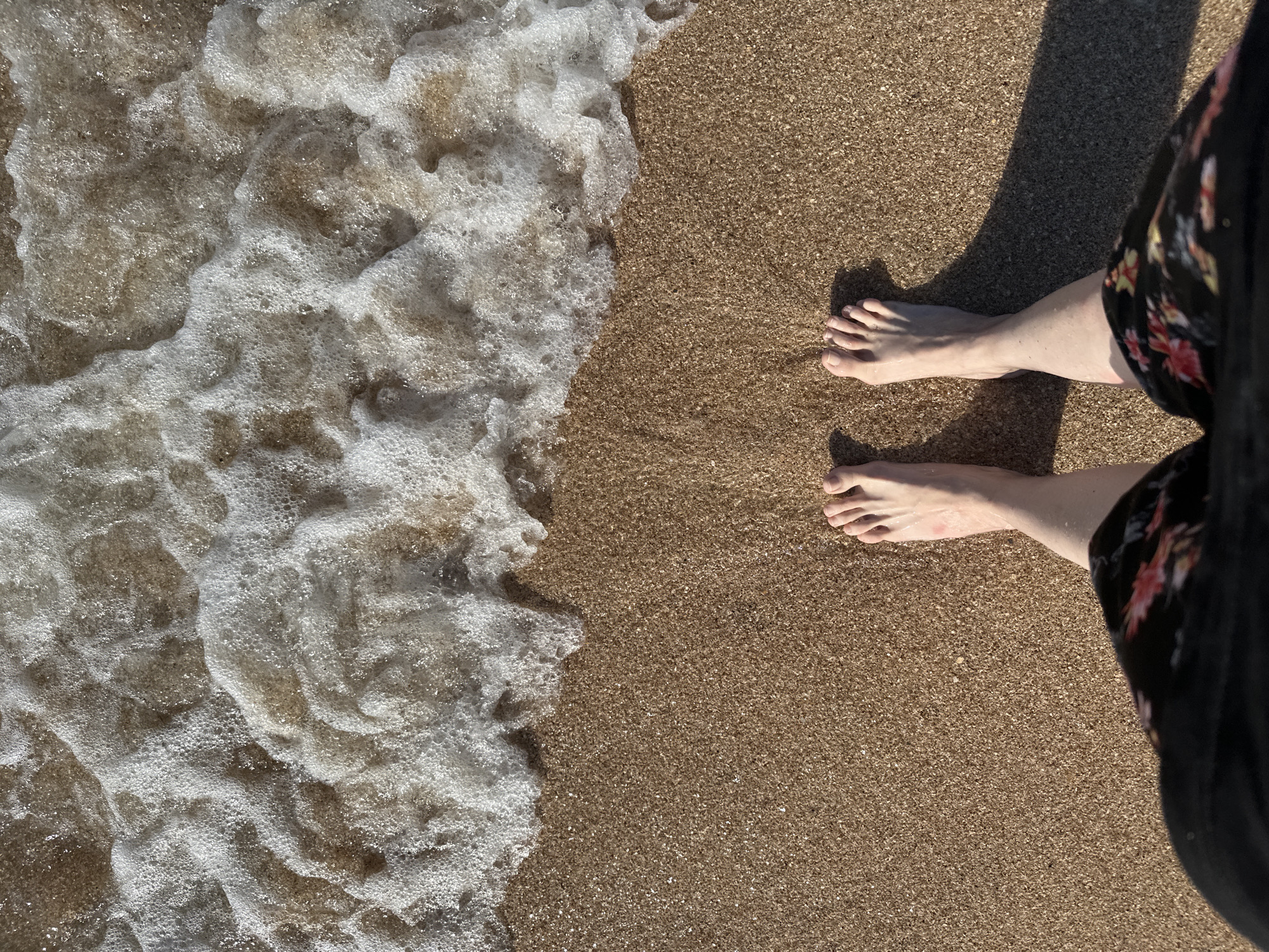 A first-person perspective shot shows a person's bare feet standing on a wet, sandy beach. The wave foam of the sea is visible in the upper part of the frame, and the sand has a fine-grained texture, with some pebbles visible. The person is wearing a black skirt with a floral pattern. The scene is in Golubitskaya, Russia.