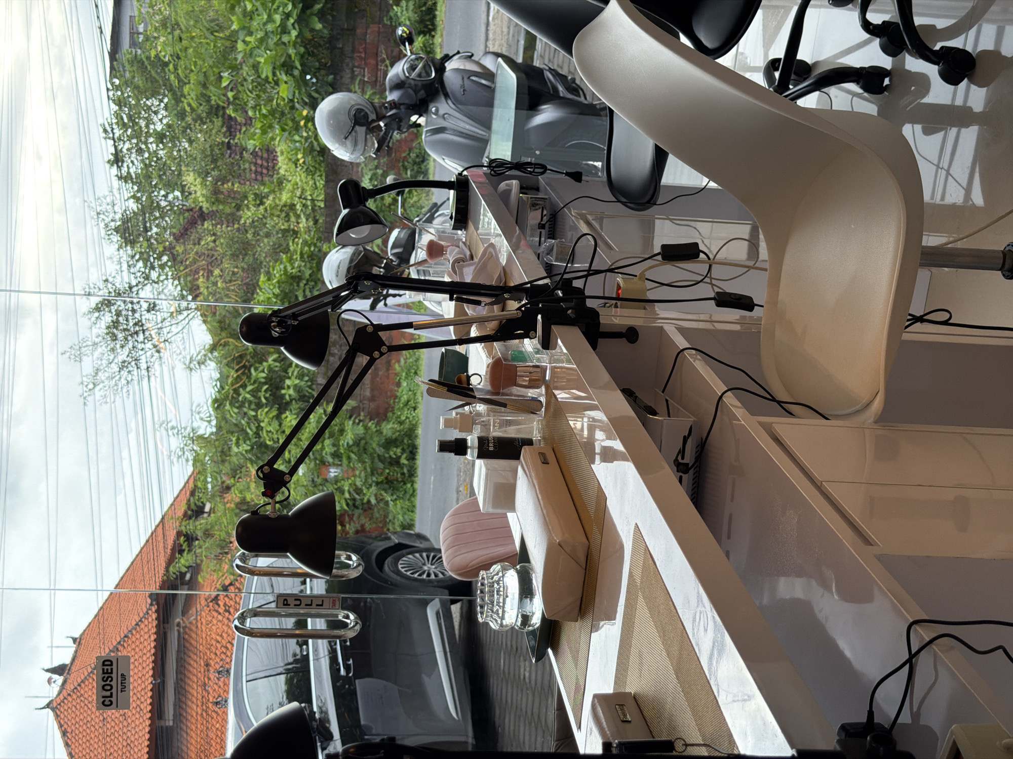 The image shows a nail salon in North Kuta, Indonesia. The salon has multiple manicure stations with adjustable black lamps. Various nail care products and tools are arranged on the white countertops. Through the glass window, the exterior reveals a scooter parked on the street, lush greenery, and a building with a red-tiled roof. A "CLOSED" sign is visible on the window.