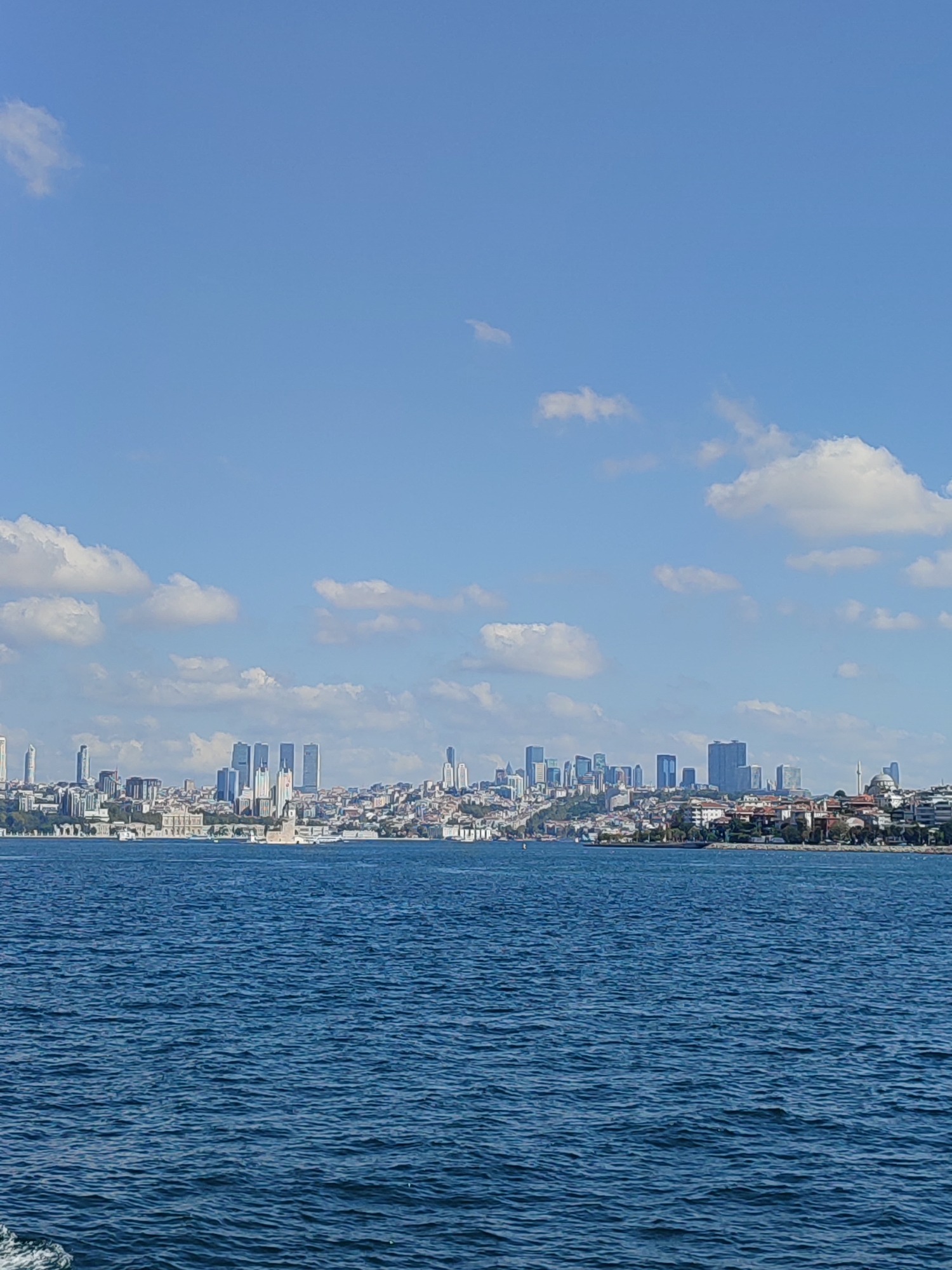 The image shows a panoramic view of the Üsküdar district of Istanbul, Turkey, from across the Bosphorus Strait. The foreground is dominated by deep blue water with gentle ripples. In the background, a dense urban landscape stretches along the coastline under a bright blue sky dotted with white, fluffy clouds.
The skyline is characterized by a mix of modern skyscrapers and older, lower-rise buildings. Prominent among the skyscrapers are several tall, rectangular buildings clustered together. To the right of these, a minaret of a mosque rises above the other structures. Red-tiled roofs are visible on many of the buildings, suggesting a mix of residential and commercial properties. Dense greenery can be seen interspersed among the buildings, particularly on the hillsides. The overall impression is of a bustling city viewed from a distance, with the calm waters of the strait separating the viewer from the urban expanse. There are no people or vehicles clearly visible in the image.