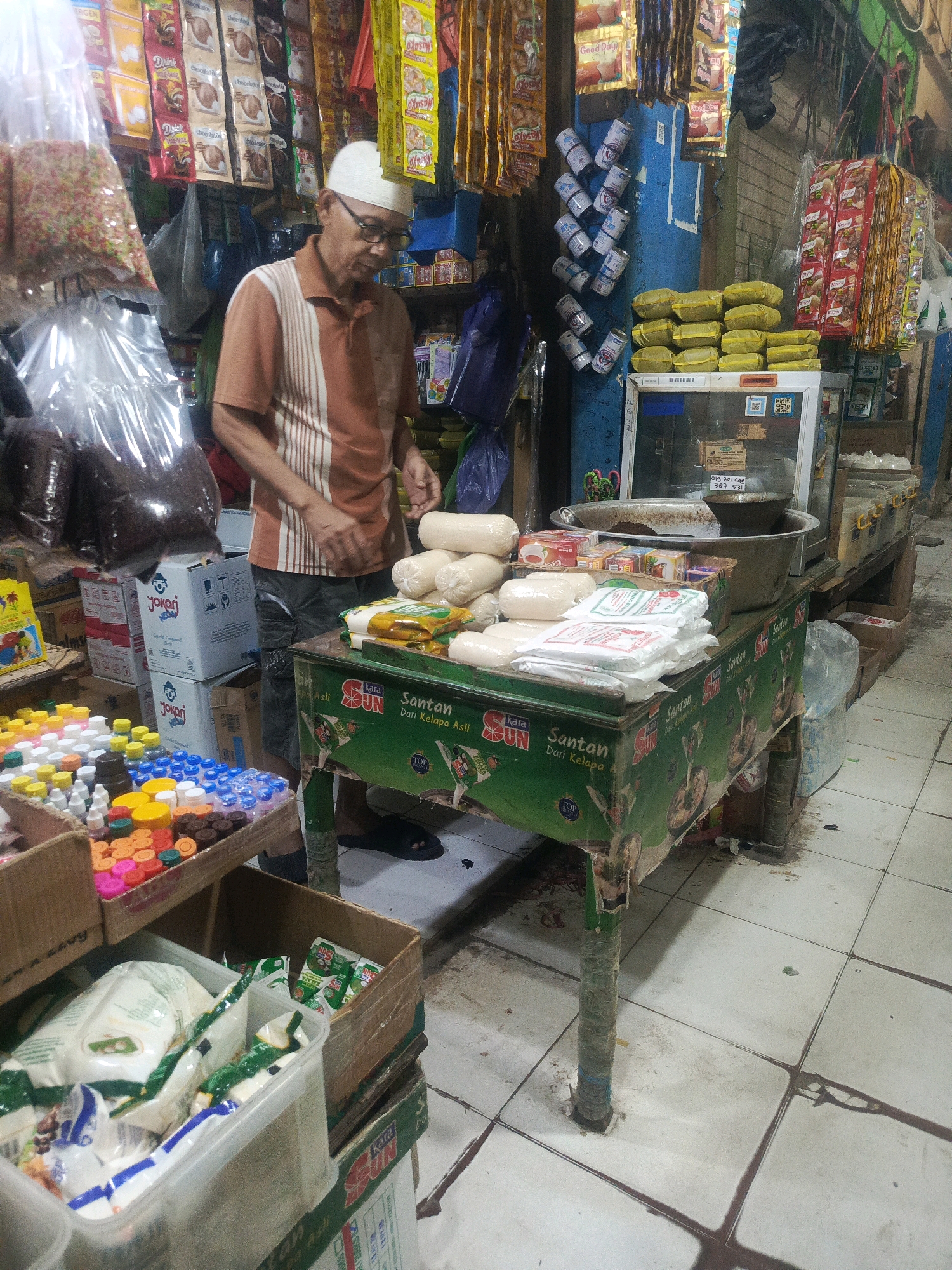 A man wearing glasses and a white cap is standing in a market stall in Kendari, Indonesia. He is wearing a short-sleeved orange and white striped shirt and dark shorts. His hands are visible, and he appears to be arranging or handling items on a green table in front of him. The table is covered with a green cloth featuring text and images of what appears to be coconut milk.
The stall is filled with various goods. In the foreground, there are boxes containing packaged food items, including bags of rice or flour and smaller packets. To the left of the man, there are several bottles and containers of colorful liquids, likely food coloring or flavorings. Behind him, shelves are stocked with bags of spices or grains, and various packaged food items hang from above. A large, clear plastic bag filled with what looks like sprinkles or small candies is also visible.
The environment appears to be an indoor market, with tiled floors that are somewhat dirty. The lighting is dim, suggesting it might be an enclosed space or an area with limited natural light. The overall impression is of a bustling, well-stocked market stall.