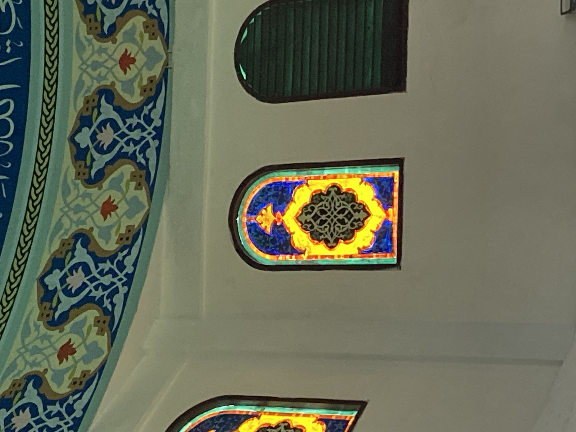 The image shows a close-up view of the interior of a building, likely a mosque, with ornate architectural details. The ceiling features intricate blue and green patterns with floral motifs, characteristic of Islamic art. Below the ceiling, there are arched windows with stained glass. One prominent stained-glass window displays a vibrant geometric design in shades of blue, yellow, and red. To its right, another arched opening is covered with dark green louvered shutters. To the left, a partial view of another stained-glass window is visible, also featuring bright colors. The walls are painted white and appear to be smooth, creating a contrast with the decorative elements. The lighting suggests the scene is illuminated by ambient light, possibly filtering through the stained glass. Based on the architectural style and decorative elements, the image is likely from a mosque in Lhokseumawe, Indonesia, known for its Islamic heritage and architecture.