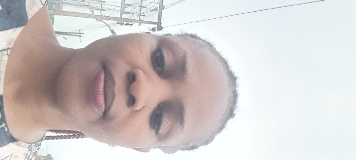 An adult female, characterized by a medium-dark complexion and braided hair, is depicted in a close-up, head-and-shoulders view. Her dark eyes are directed towards the viewer, and her full lips maintain a neutral expression. She is wearing a patterned garment featuring light and dark tones. The outdoor background consists of an overcast, light grey sky. To the left, a utility pole structure with multiple power lines and a transformer is clearly visible, characteristic of urban infrastructure. Additional utility poles and faint distant structures are present further back. The scene is located in Aba, Abia, Nigeria.