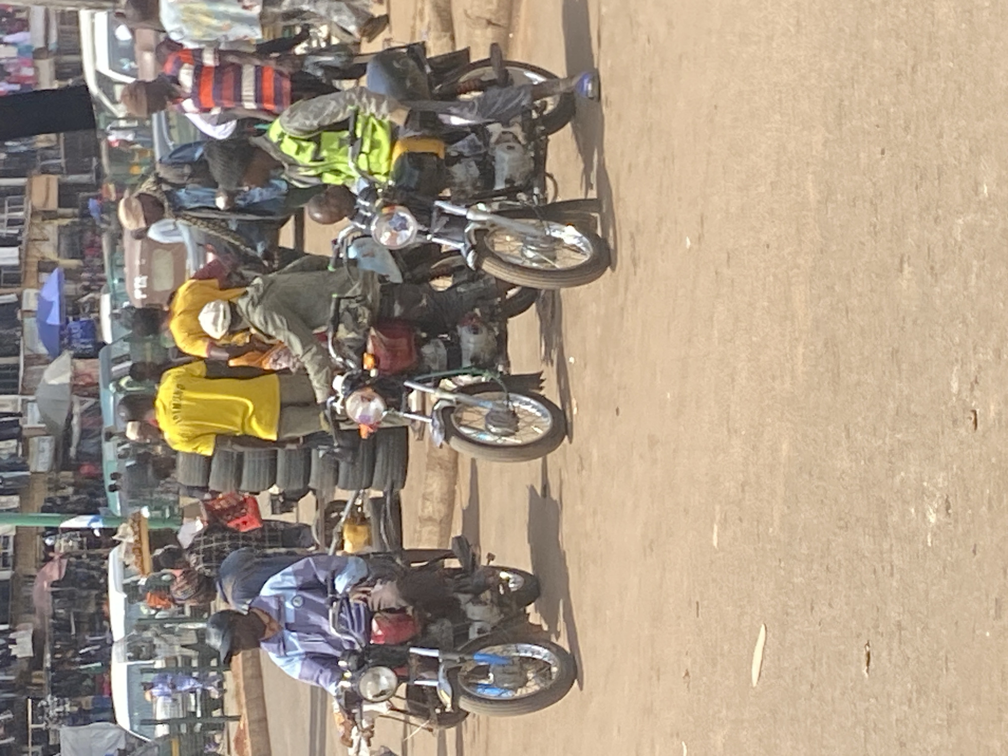 The image captures a bustling street scene in Bukuru, Nigeria, under bright, clear daylight. The setting appears to be a market area or a busy thoroughfare, characterized by numerous people and motorcycles. The ground is a light-colored, dusty surface, with visible shadows indicating a sunny and dry day.
The foreground and midground are filled with multiple motorcycles, likely operating as "okadas" or motorcycle taxis. Several individuals, primarily men, are either riding, sitting on, or standing near these bikes. Some motorcycles are visibly loaded with goods, such as stacked tires and various packages, suggesting their use for both passenger and cargo transport. People are dressed in casual attire, including shirts and trousers, with some wearing caps. On the back of a prominent yellow shirt, the word "DIMUNO" is partially visible.
In the background on the left, there are hints of market activity, including stalls or shops displaying various goods, and a blue umbrella providing shade. Partially visible cars or vans further down the street indicate a mixed traffic environment. The scene portrays the daily commerce and transportation activities typical of a vibrant West African street.