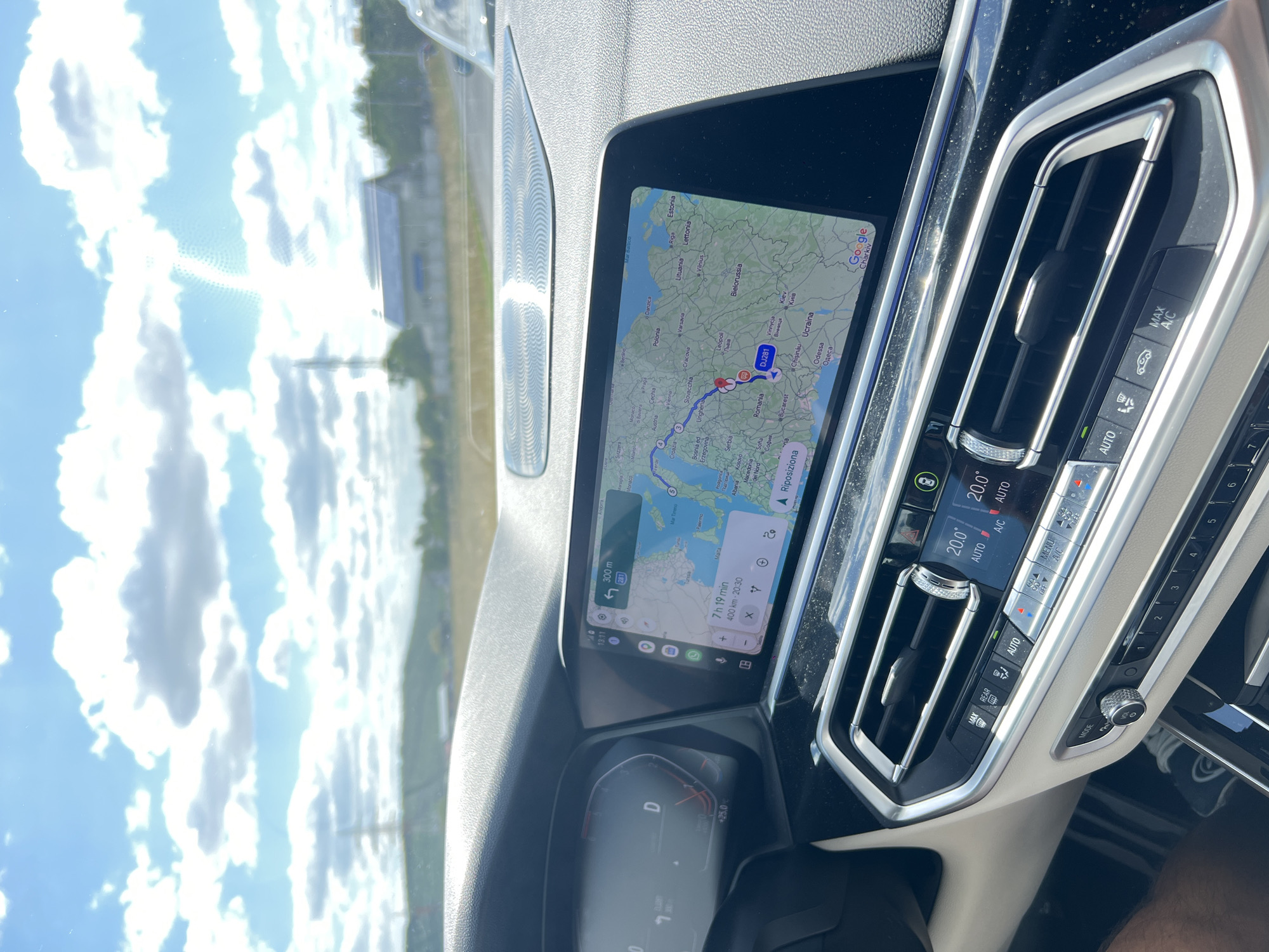 The image shows the interior of a car with a navigation screen displaying a map of Europe. The route appears to be traveling from a location in Eastern Europe towards Western Europe. The screen shows the current time as 13:11 and the remaining travel time as 7 hours and 19 minutes for a distance of 400 kilometers. A red pin is visible on the map, marking the city of Podu Iloaiei, Romania. The dashboard also shows the car's speed is 0 km/h and the outside temperature is 20.0 degrees Celsius, with the climate control set to AUTO. The car is stationary, as indicated by the speed. The environment outside the car is sunny with a partly cloudy sky.