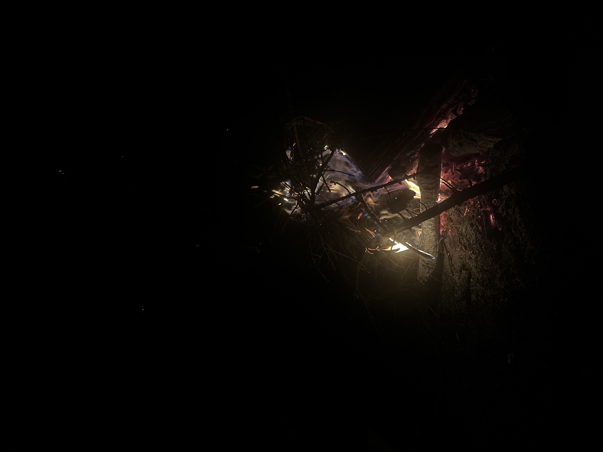 The image depicts a campfire in a dark environment. The fire is contained within what appears to be a natural or man-made fire pit, surrounded by earth and logs. Flames and glowing embers are visible, illuminating the immediate area around the fire. Thin branches and other smaller pieces of wood are burning. The scene is predominantly dark, with the fire acting as the main light source. No people are visible. The location is Юзуфоўскі сельскі Савет, Belarus.