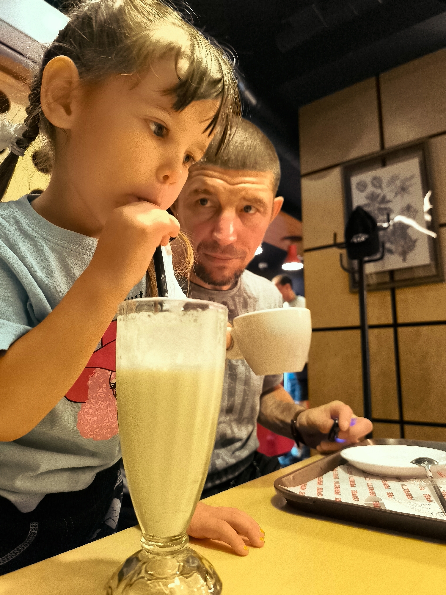 A young girl with braided hair is drinking a milkshake through a straw. She is wearing a light blue t-shirt. Beside her, a man with short hair is holding a white cup and looking towards the camera. The scene appears to be in a cafe or restaurant, suggested by the table, tray, and framed pictures on the wall. The lighting is warm and atmospheric, with some background elements suggesting this location is in Karaganda, Kazakhstan.