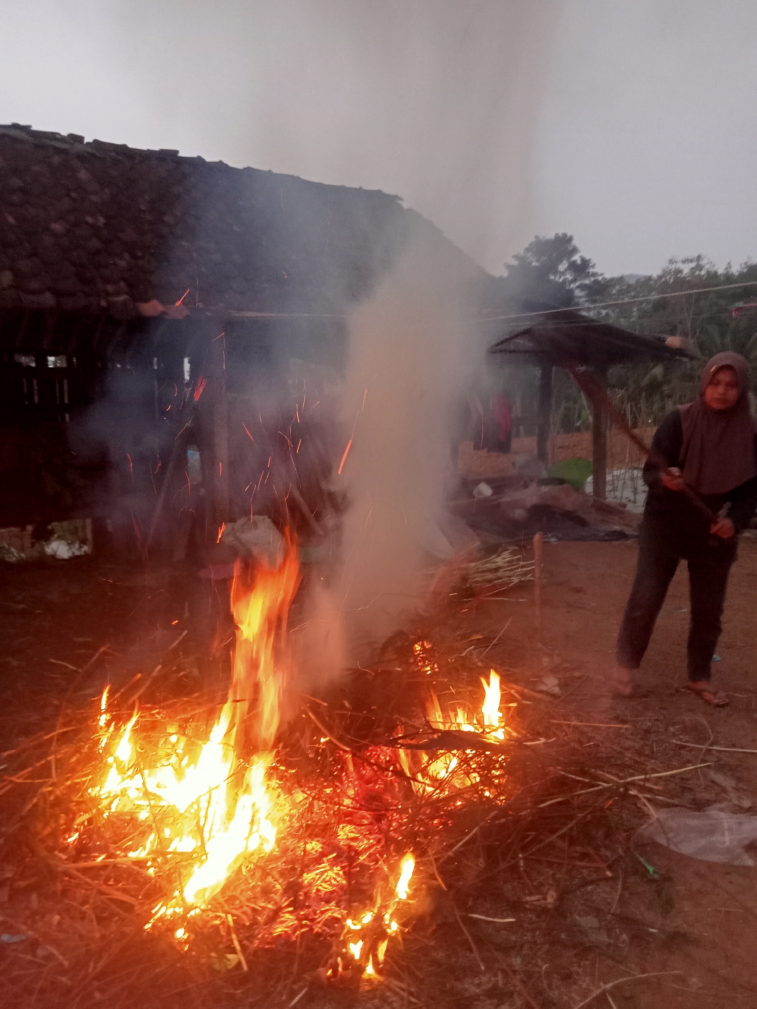 An outdoor scene, possibly in Tanggaran, Indonesia, depicts a fire burning in front of a rustic structure with a thatched roof. A person wearing a hijab and dark clothing stands to the right of the fire, holding a stick. Smoke rises from the fire, obscuring some of the background elements, including additional structures and vegetation. The ground is covered in dry debris, likely the fuel for the fire. The lighting suggests it may be dawn or dusk.