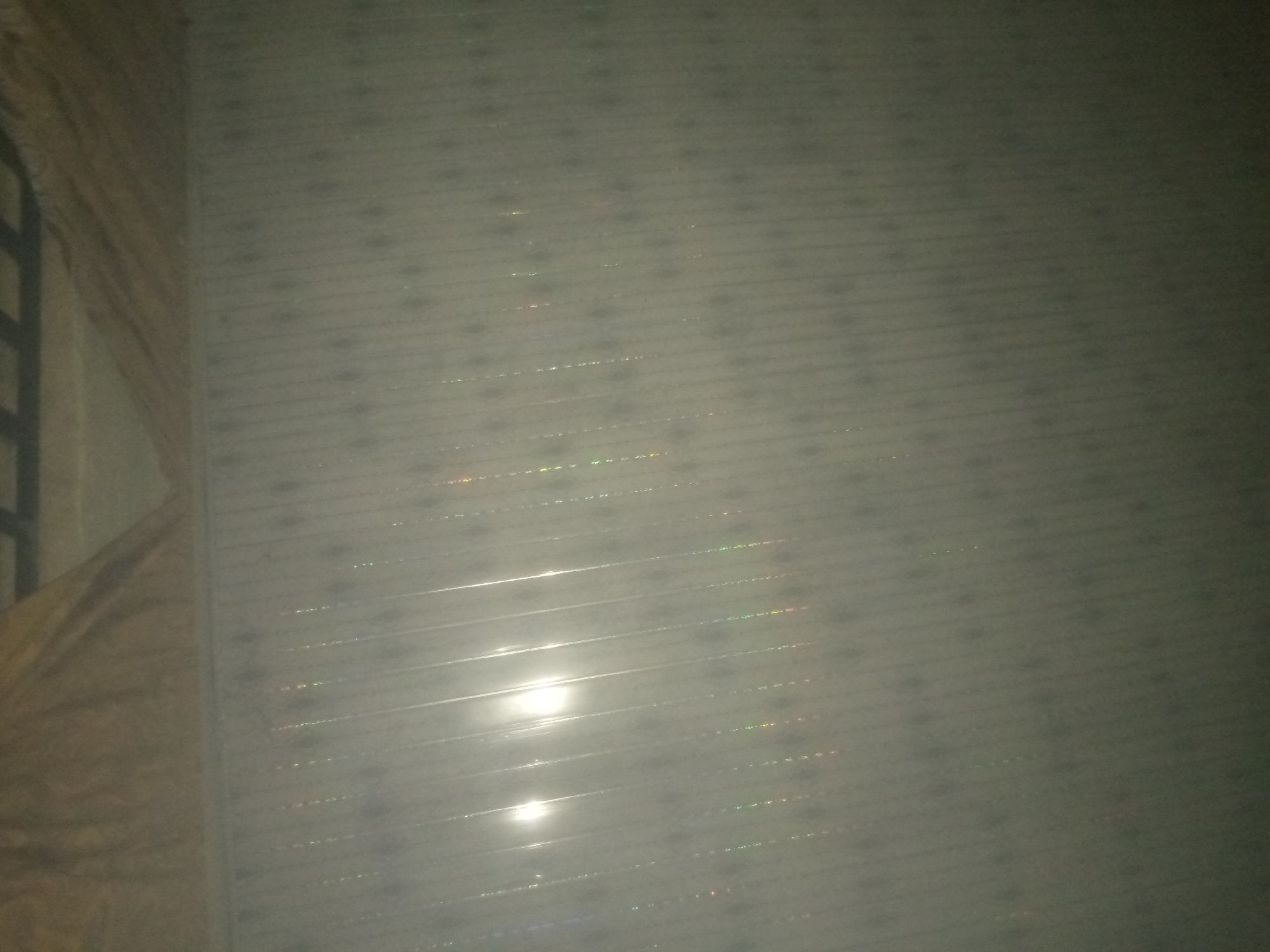 The image depicts a close-up, horizontal view of a textured surface, likely a wall or ceiling, with a repeating pattern of horizontal lines. Small, reflective or iridescent flecks are scattered across the surface, catching the light. A bright, white light source is reflected strongly in the lower central portion of the image, creating a lens flare effect. To the left, a draped fabric, appearing to be a blanket or curtain, is visible, with a darker, possibly metal, bar-like structure partially obscuring it at the top left. The overall lighting is dim, suggesting an indoor or low-light environment. The location is identified as Jauro Yinu, Nigeria.