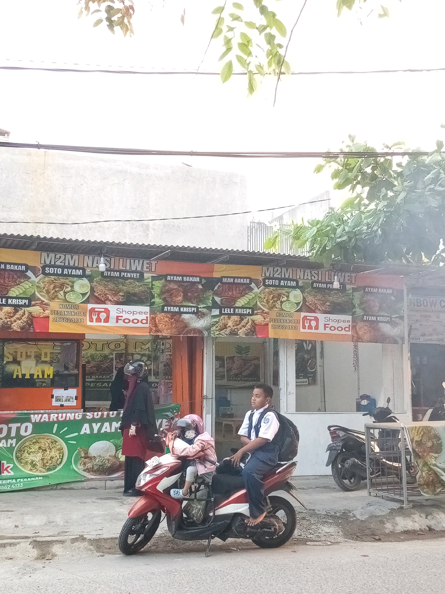 In Mangunjaya, Indonesia, a street scene unfolds with a red and white motorbike parked on the side of a road. A young boy wearing a white collared shirt, a blue vest, dark pants, and a backpack is seated on the motorbike, his bare feet resting on the engine casing. He looks to his left. To the left of the boy, another motorbike is partially visible. A woman wearing a dark hijab and a red outer garment stands behind the motorbike the boy is on, facing to her right. Further to the left, a sign for "M2M NASI LIWET" is prominently displayed, showcasing various Indonesian dishes like "SOTO AYAM" and "AYAM PENYET." The sign also features logos for "Shopee Food" and a phone number. Adjacent to the sign is a shop counter with various food items. In the background, a light-colored building stretches across the frame, with some greenery overhead. Another motorbike is parked on the far right, near a metal cage displaying food. The overall environment appears to be a commercial street.