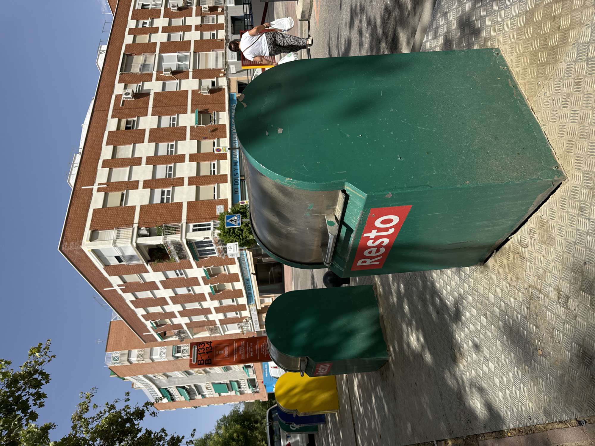 In the city of Murcia, Spain, a row of colorful recycling bins stands on a metal-plated sidewalk. The bins are predominantly green, with one yellow and one blue bin visible further down the line. The front-most green bin has a red rectangle with the word "Resto" in white lettering. Behind the bins, a modern building with a red brick facade and white checkered patterns rises, featuring balconies with white railings and green awnings. A sidewalk with a distinctive diamond plate pattern runs in front of the bins. Sunlight casts shadows on the sidewalk and bins. In the background, a woman walks away from the camera, carrying two white plastic bags. She is wearing patterned trousers and a white top. A signpost with orange and white text is visible near the bins. The sky is clear and blue.