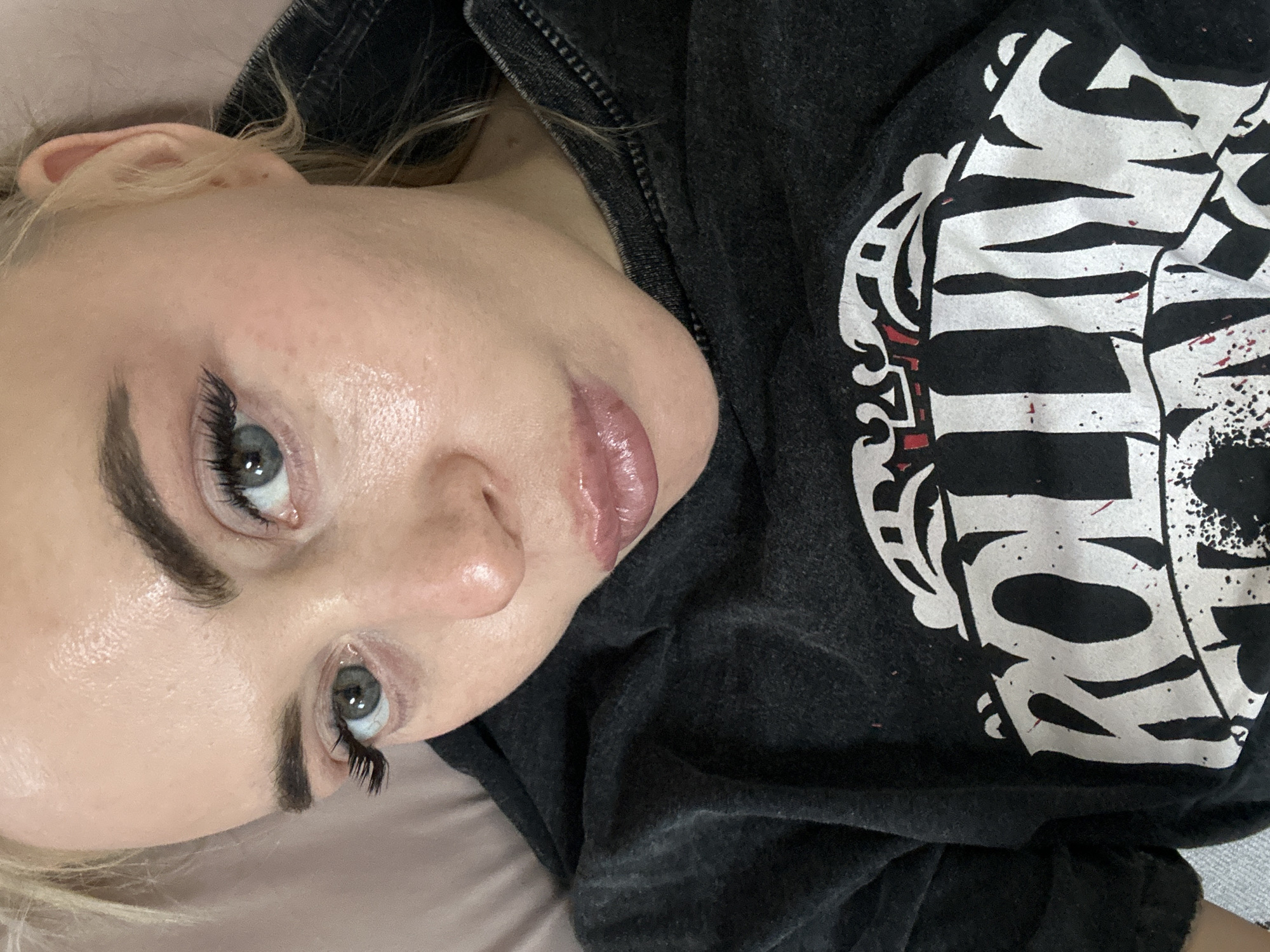 A close-up, angled shot features a fair-skinned individual with light blonde hair, light eyes, and full, glossy lips. Their makeup includes dark eyebrows and enhanced eyelashes. They are wearing a dark grey hooded sweatshirt. The sweatshirt has a white graphic on the front. The background appears to be a light pink or beige surface, possibly bedding or a pillow. There is no information available in the image about the city of Высокий, Russia.