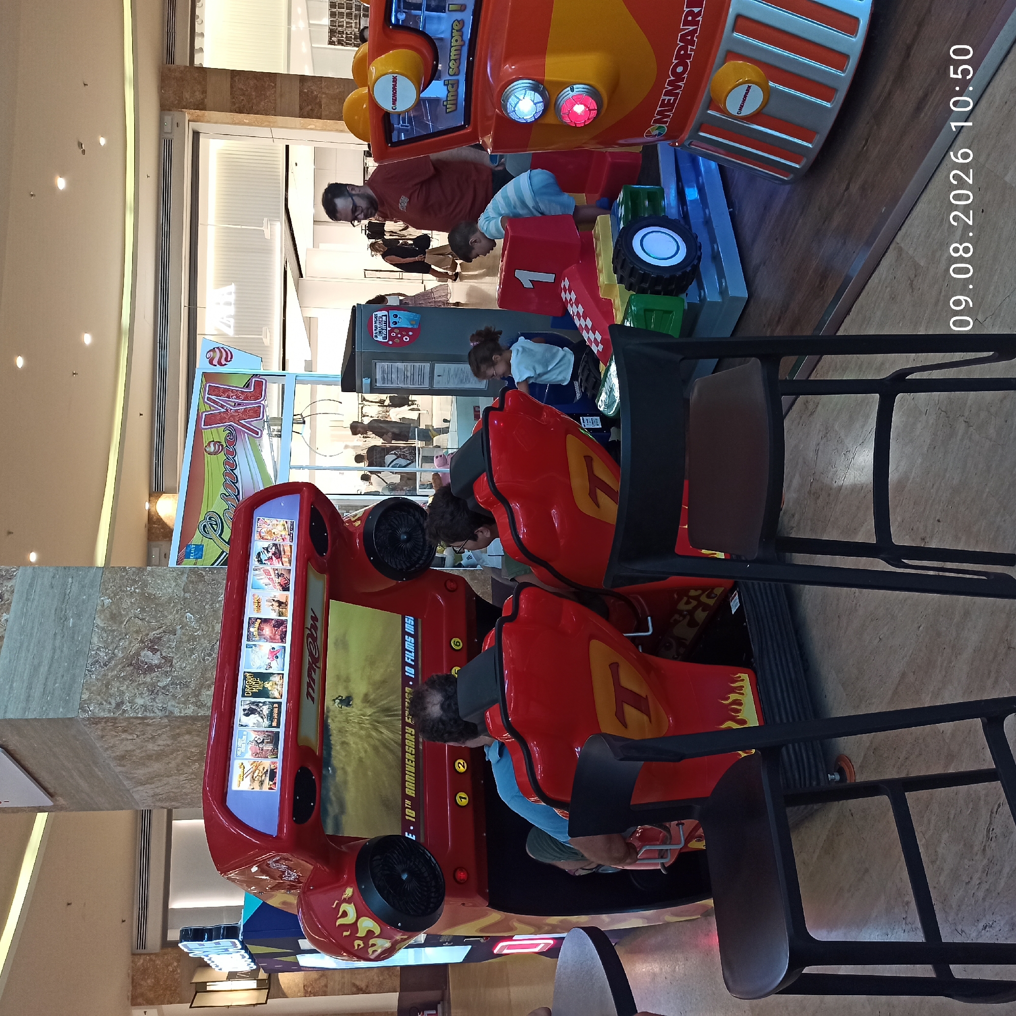 This image captures a lively scene inside a shopping mall, possibly in Molfetta, Italy. The foreground features two red bar stools, one of which is closer to the viewer, with a dark brown seat. Behind these stools, a bright red racing arcade game is prominently displayed, its screen showing a driving simulation. The game unit has flame decals on its sides and is designed to seat two players. One player, a man with dark, curly hair, is seated in the game. To the right of this game, another smaller ride-on toy, resembling a race car, is occupied by a young boy wearing a striped shirt and blue shorts. A young girl with glasses, wearing a light blue shirt, is also interacting with this ride. A man with a beard and glasses, wearing a red shirt, stands near the race car ride, observing the children. In the background, more arcade games and store displays are visible, including a large sign that reads "Cosmic XL." Shoppers are seen in the distance, and the mall's architecture includes marble-patterned columns and a high, illuminated ceiling. The overall atmosphere suggests a recreational area within a commercial setting.