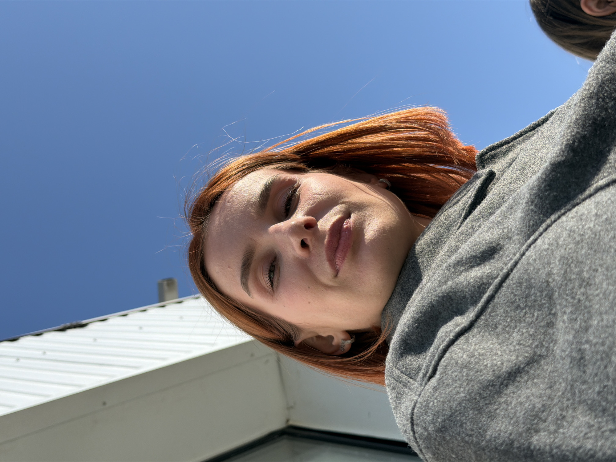 A low-angle shot of a young woman with auburn hair, wearing a gray coat or jacket, against a blue sky background. A white building structure with a corrugated metal roof is visible in the lower left of the frame. Another person's ear is visible in the upper right of the frame. The location is not discernible from the image. No specific information about the city of Chaikovsky, Russia, is evident.