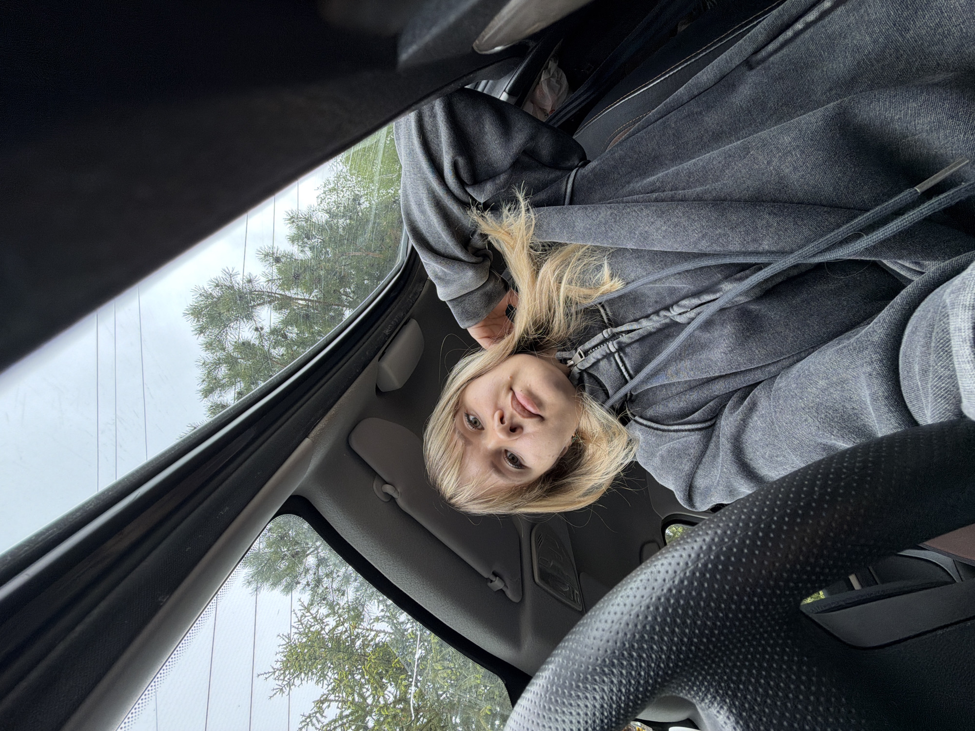 A young woman with blonde hair sits in the driver's seat of a car. She wears a grey hooded sweatshirt. The steering wheel is visible in the foreground. Through the car's windshield, trees are visible, suggesting an outdoor location. There is no specific information to determine the location is Chaikovsky, Russia.