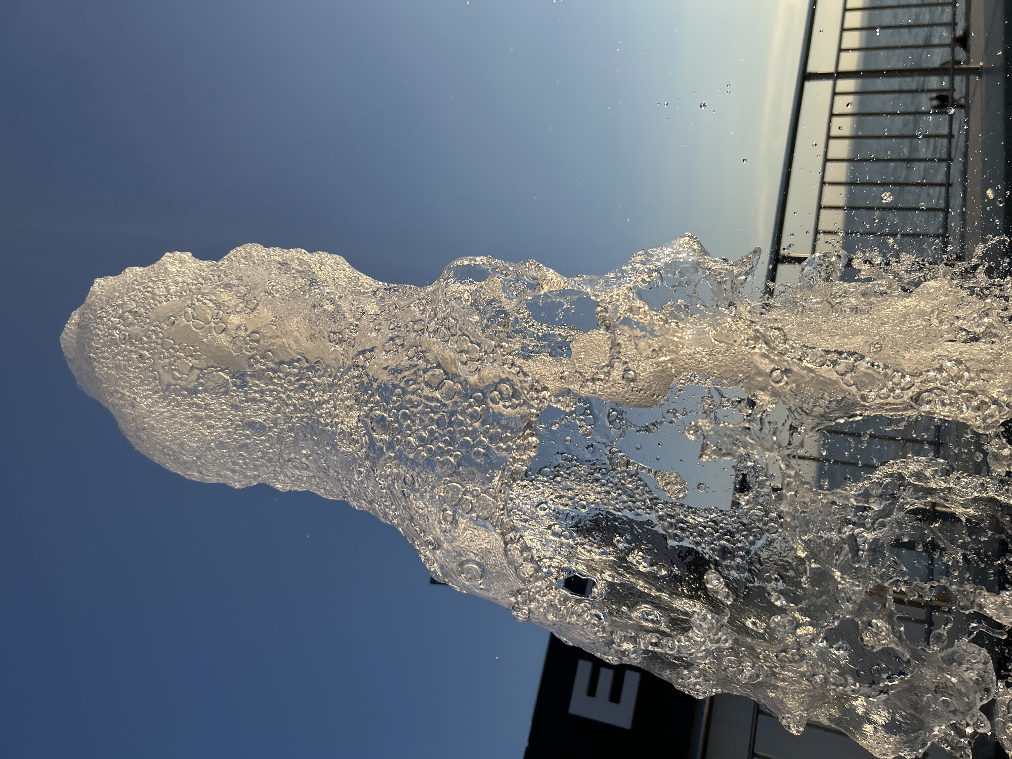 The image captures a dynamic water fountain against a clear blue sky. The water is frozen in motion, forming an abstract shape with numerous bubbles and droplets. In the background, a black structure with the letter "E" visible is partially obscured by the fountain's spray. Beyond that, a metal railing runs horizontally, separating the viewer from a body of water, likely the Volga River, with a faint silhouette of people visible on the embankment. The scene appears to be located in Tolyatti, Russia, possibly along its waterfront promenade.