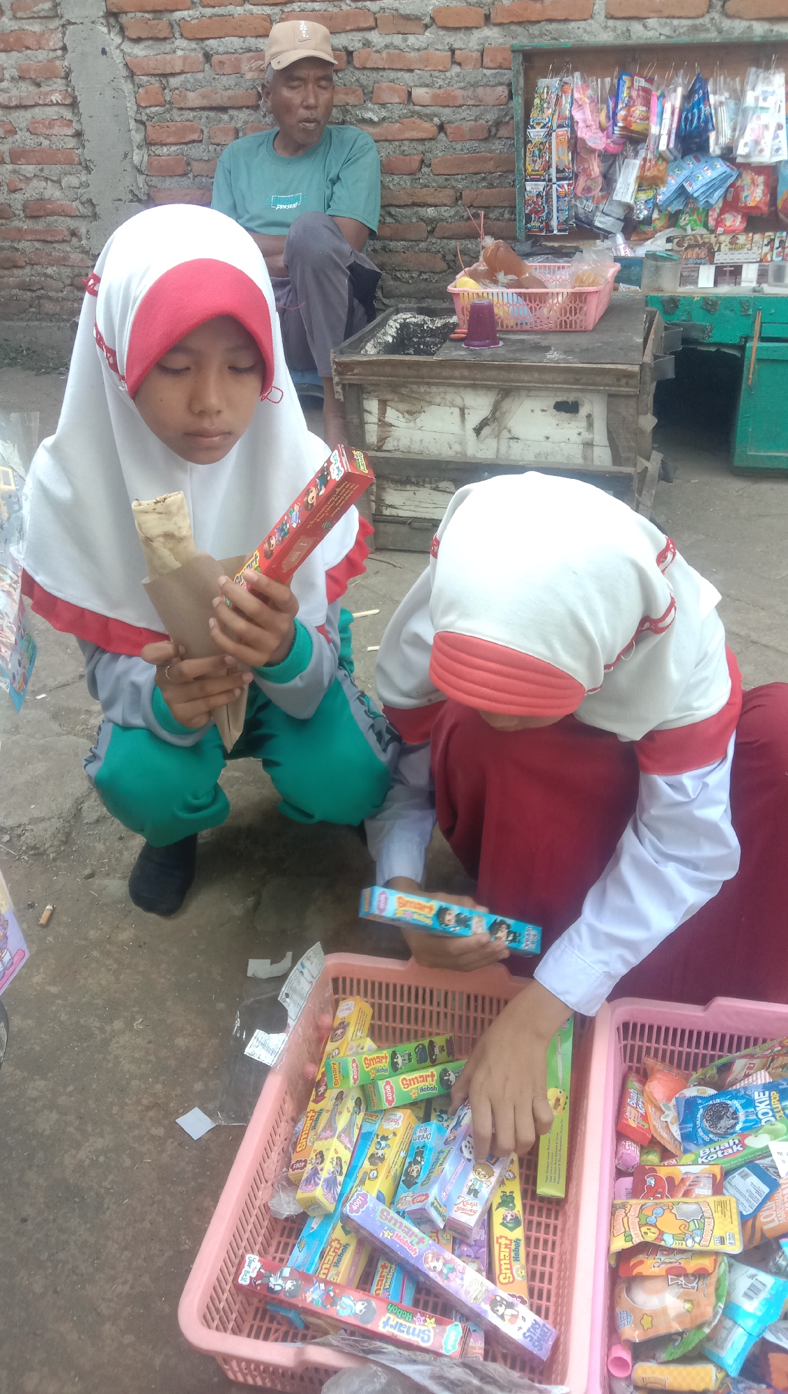 Two young girls wearing hijabs, one in white with red trim and the other in red, are squatting near a stall. The girl on the left is holding a wrapped food item and a box of candy. The girl on the right is reaching into a pink basket filled with various candies and snacks. Both girls are looking down at the items. In the background, an elderly man wearing a cap and a teal t-shirt is sitting and appears to be observing. The stall behind them is stocked with an assortment of colorful packaged goods, including candies, cookies, and possibly toys. The ground is paved with concrete, and a brick wall forms the backdrop. The scene appears to be an outdoor marketplace or a street-side stall. This scene is in Karang Anyar, Indonesia.