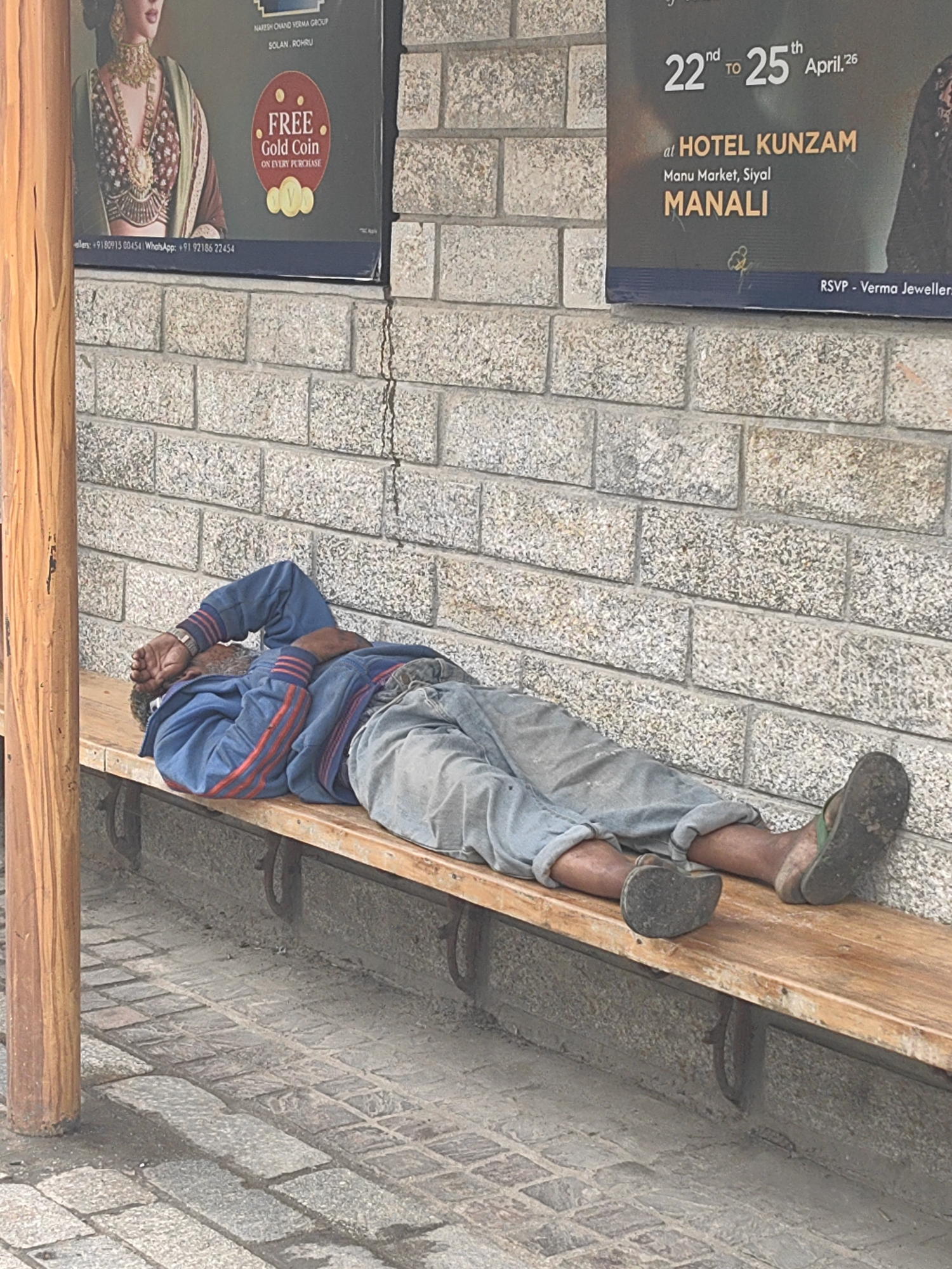 A man lies asleep on a wooden bench in front of a brick wall in Manali, India. He is wearing a blue jacket with red stripes on the sleeves and light-colored pants. His right arm is covering his face, and his left hand is resting on his chest. His legs are stretched out on the bench, and he is wearing sandals. The background shows posters on the wall, one advertising a jewelry store with a "Free Gold Coin" offer, and another for "Hotel Kunzam" in Manu Market, Siyal, Manali. The ground is paved with cobblestones.