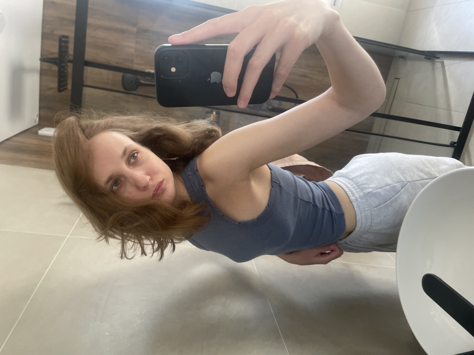 A woman with auburn hair is taking a selfie in a bathroom. She is holding a black iPhone with an Apple logo in her right hand. She is wearing a grey tank top and light grey sweatpants. The bathroom has beige tile flooring and a shelf with a black metal frame. The mirror reflects the scene. There is a white toilet in the lower right corner of the image. The location in the city of Подшивалово, Russia is not discernible from this image.