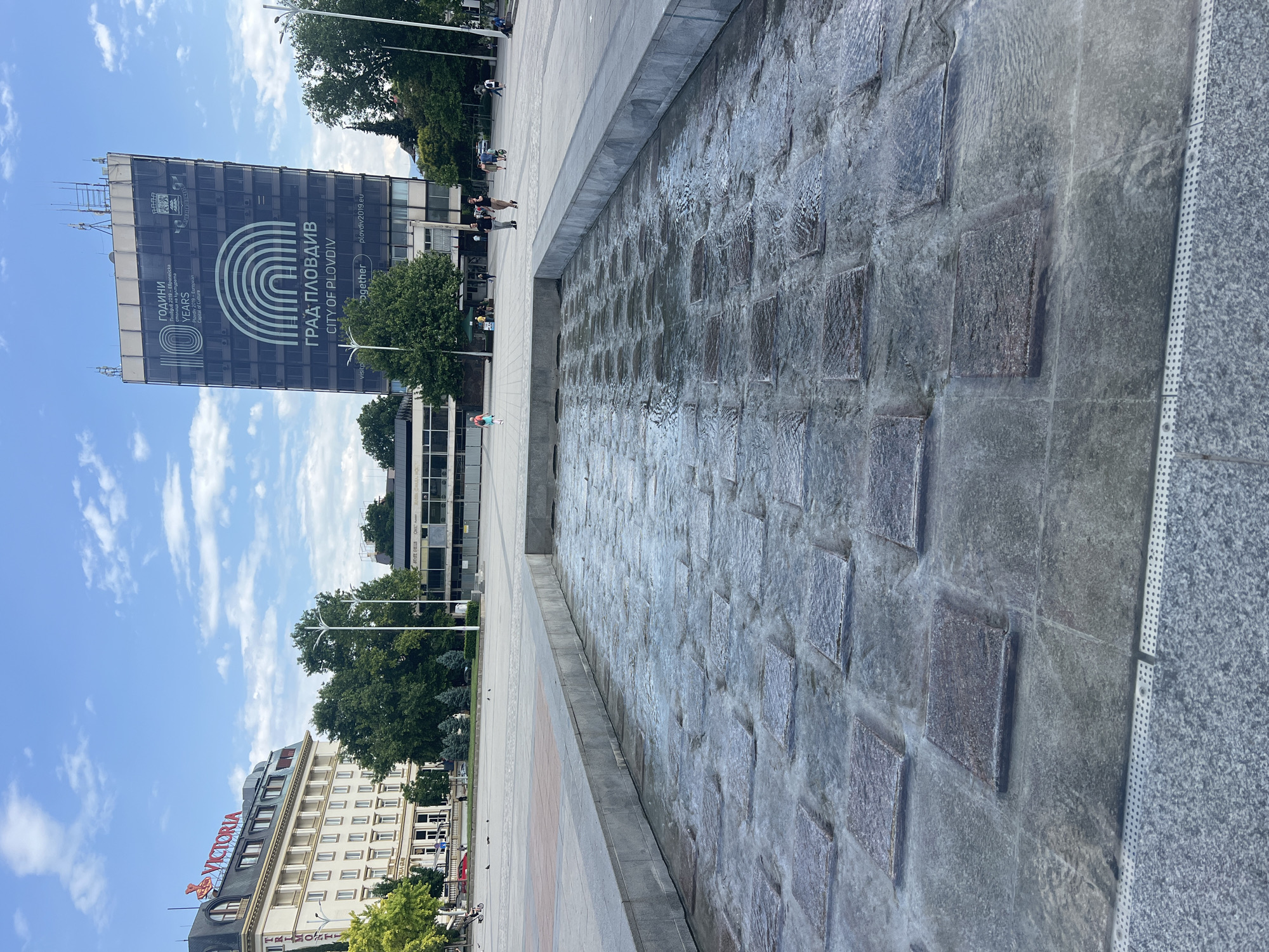 The image captures a ground-level view of a water feature located in Plovdiv, Bulgaria. The water flows over a patterned surface of square stones within a concrete basin. A paved walkway borders the water feature, offering a space for pedestrians. In the background, a tall building displays the inscription "ГРАД ПЛОВДИВ CITY OF PLOVDIV" along with "YEARS" and "Пловдив 2019 - Европейска столица на", which indicates Plovdiv's status as a European Capital of Culture in 2019. Another building with the name "VICTORIA" in red letters is also visible, with a hotel also in the image. The scene is set on a partly cloudy day with blue skies.