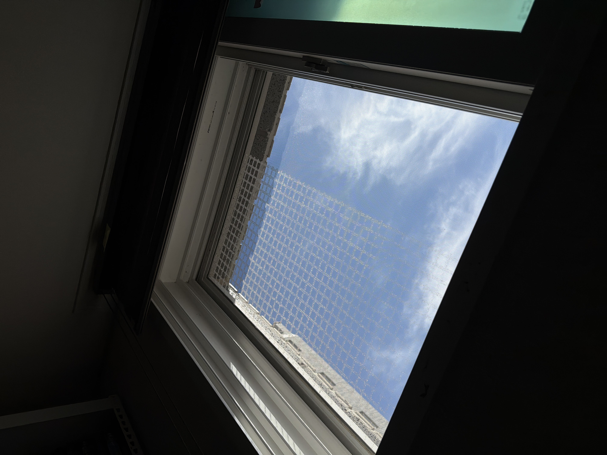 A tilted shot looks out a window onto a bright blue sky with wispy clouds. A building with a textured facade is visible across the window, partially obscured by a mesh screen. The window frame is white and grey, and a dark curtain or blind hangs above it. The overall scene appears to be from an indoor perspective, looking outwards. The image is believed to be taken in Dongducheon-si, South Korea.