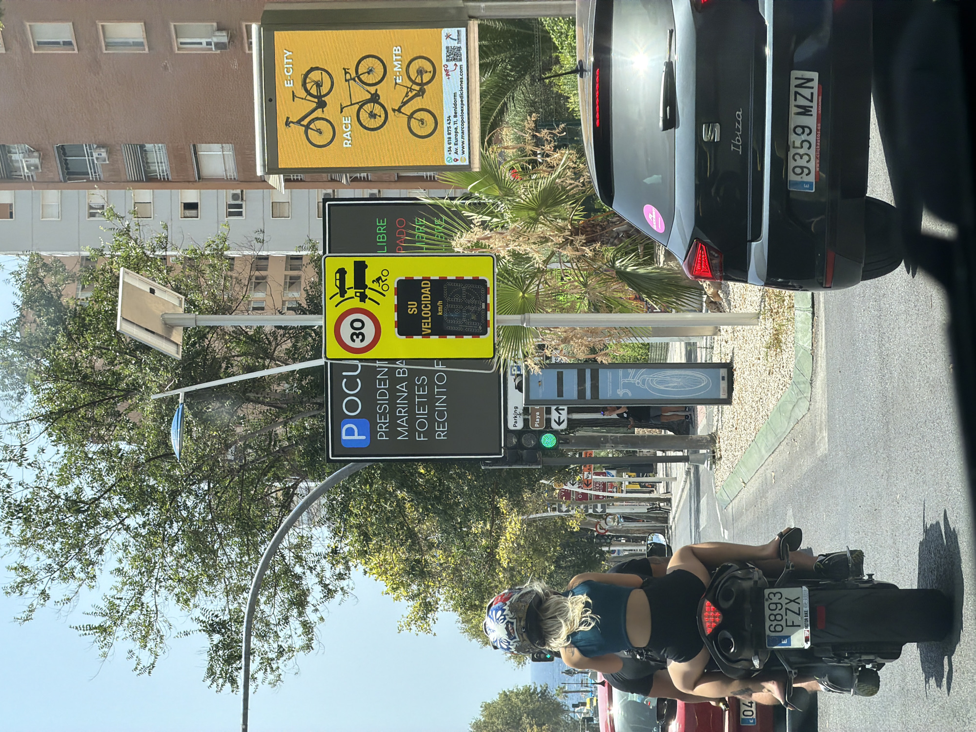 The image shows a street scene in Benidorm, Spain, on a sunny day. The foreground features the rear of a black SEAT Ibiza car, its license plate reading "E 9359 MZN". To the left, a motorcycle with two passengers is visible. The driver, a male, is partially obscured, while the female passenger, wearing a helmet, is facing away from the camera. She is wearing a teal crop top and black shorts. Her legs are visible, and she is wearing flip-flops. The motorcycle's license plate reads "E 6893 FZX".
In the background, there are street signs. One sign is yellow with black text and graphics, showing bicycles of different types: "E-CITY", "RACE", and "E-MTB". Another sign is black with white text and a blue "P" logo, likely indicating parking. There's also a speed limit sign showing "30" and a sign that says "SU VELOCIDAD" (Your Speed) with a digital display. Traffic lights are visible, with a green light illuminated. Buildings line the street, some with balconies and air conditioning units. Trees and palm trees add to the Mediterranean ambiance. The ocean is visible in the distance.