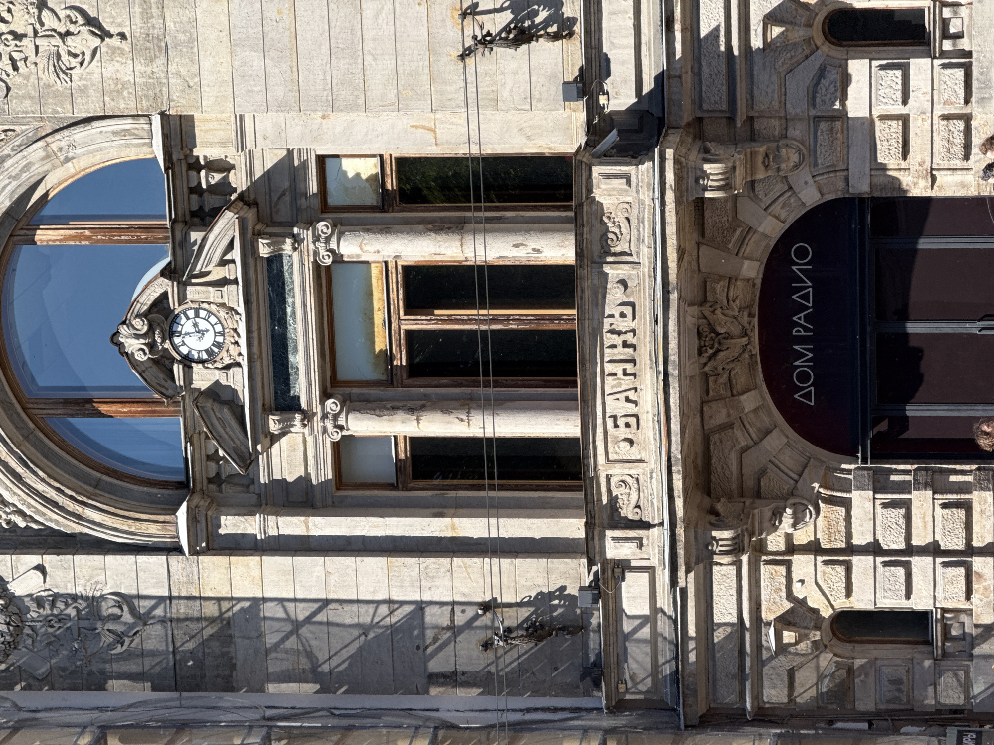The image shows the facade of an old building in Saint Petersburg, Russia. The building's architecture is ornate, with classical elements such as columns, arched windows, and decorative carvings. A large clock is visible above a balcony, and the word "БАНКЪ" (Bank) is inscribed on the balcony's facade in Cyrillic. Below the balcony, an arched entrance features the words "ДОМ РАДИО" (House of Radio) in Cyrillic. The building is constructed of light-colored stone, and shadows from surrounding structures fall across its facade. The sky is a clear blue. The image appears to be taken during the daytime. There are no people clearly visible in the image, but the presence of a doorway suggests it is an accessible building. The overall impression is one of historical grandeur and the juxtaposition of different establishments within the same building.